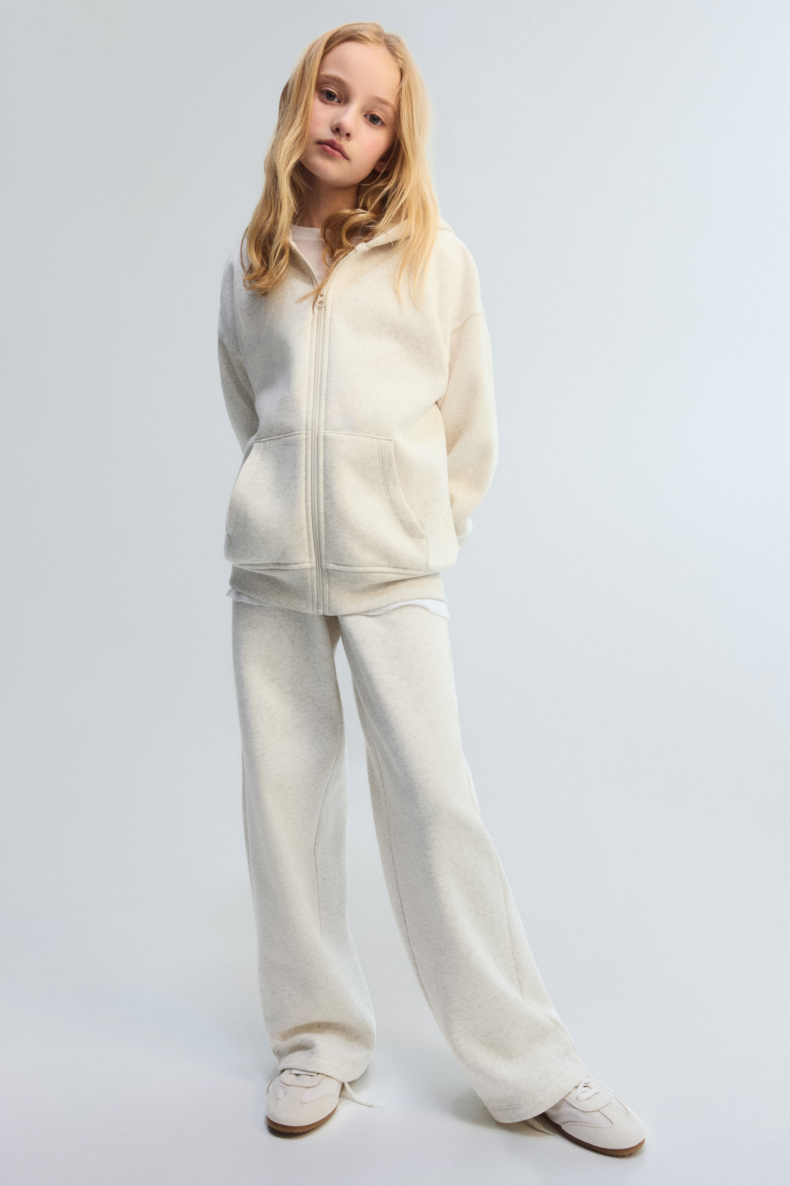A heathered light beige tracksuit is showcased, featuring a full-zip hoodie with a hood and front patch pockets, accompanied by matching wide-leg trousers. A white t-shirt is visibly layered beneath the hem of the hoodie. - 1