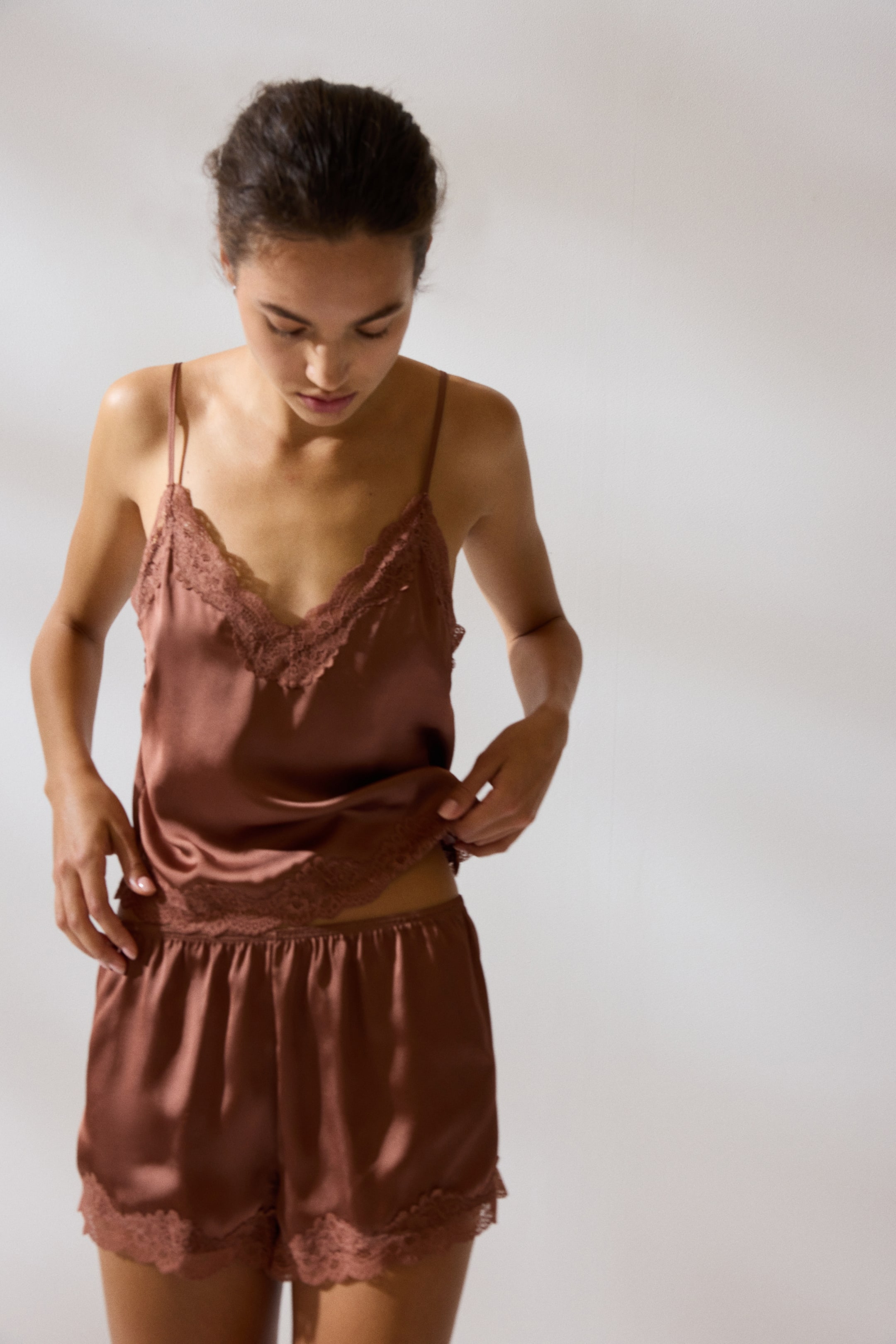 Visa större bild: En rödbrun satinpyjamas, bestående av ett linne med spagettiband och V-ringning samt matchande shorts, bärs av modellen. Både linnet och shortsen har spetsdetaljer, och modellen håller försiktigt i linnets fåll.