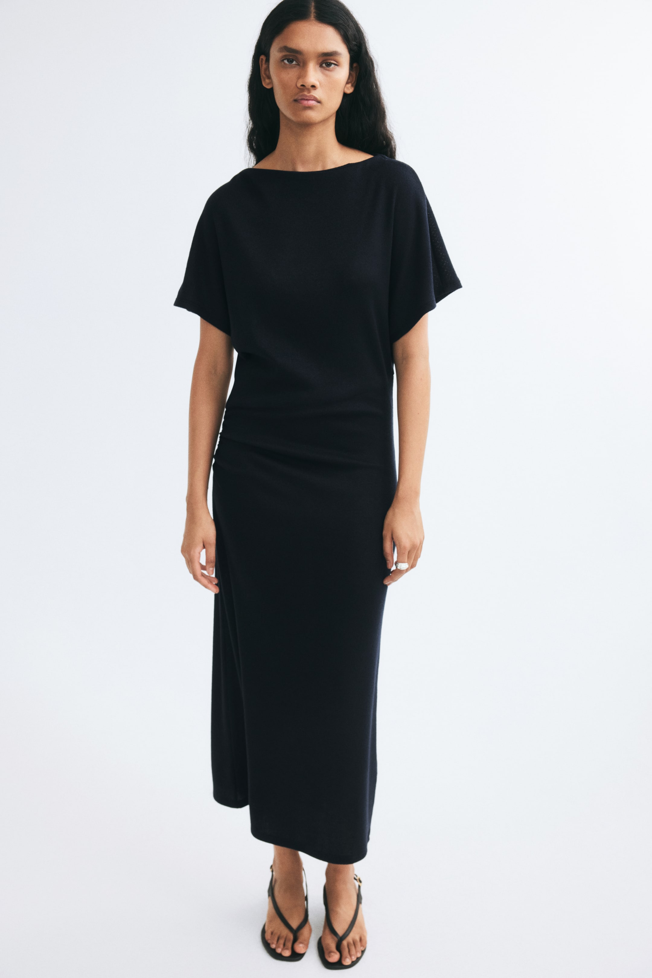 View larger image: The model is wearing a long black knit dress featuring a delicate boat neck, a relaxed-fit bodice, and short butterfly sleeves. The dress has a diagonal waist seam with gathering at the hip, flowing down to an ankle-length hem.