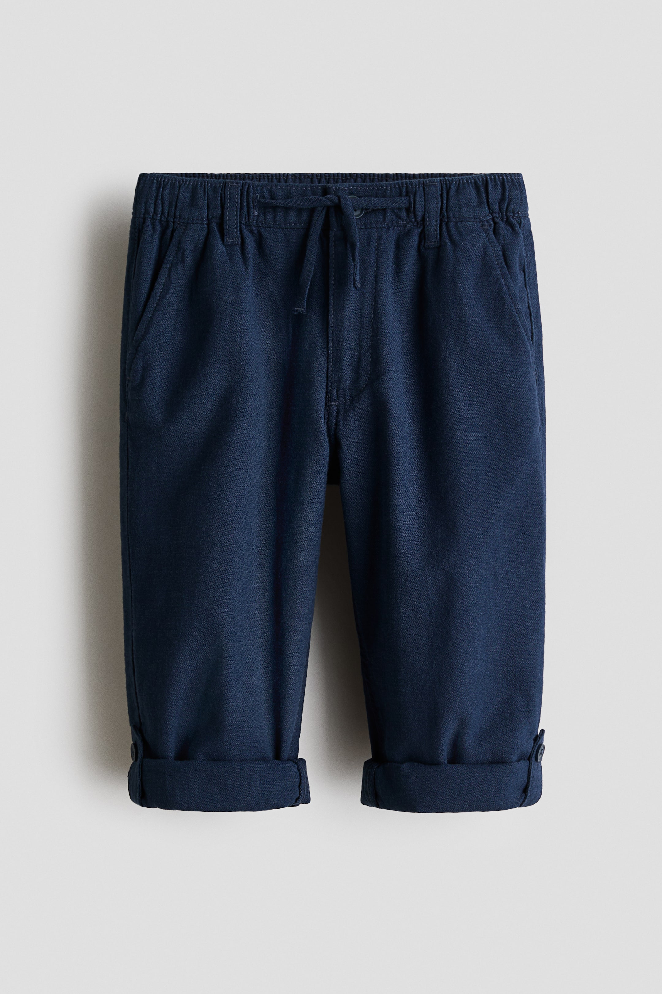 View larger image: Dark blue trousers crafted from textured cotton twill are showcased from the front, boasting an elasticated drawstring waist with a faux fly and slanted side pockets. The relaxed-fit trousers taper at the legs, which are turned up at the cuffs with a visible button tab.