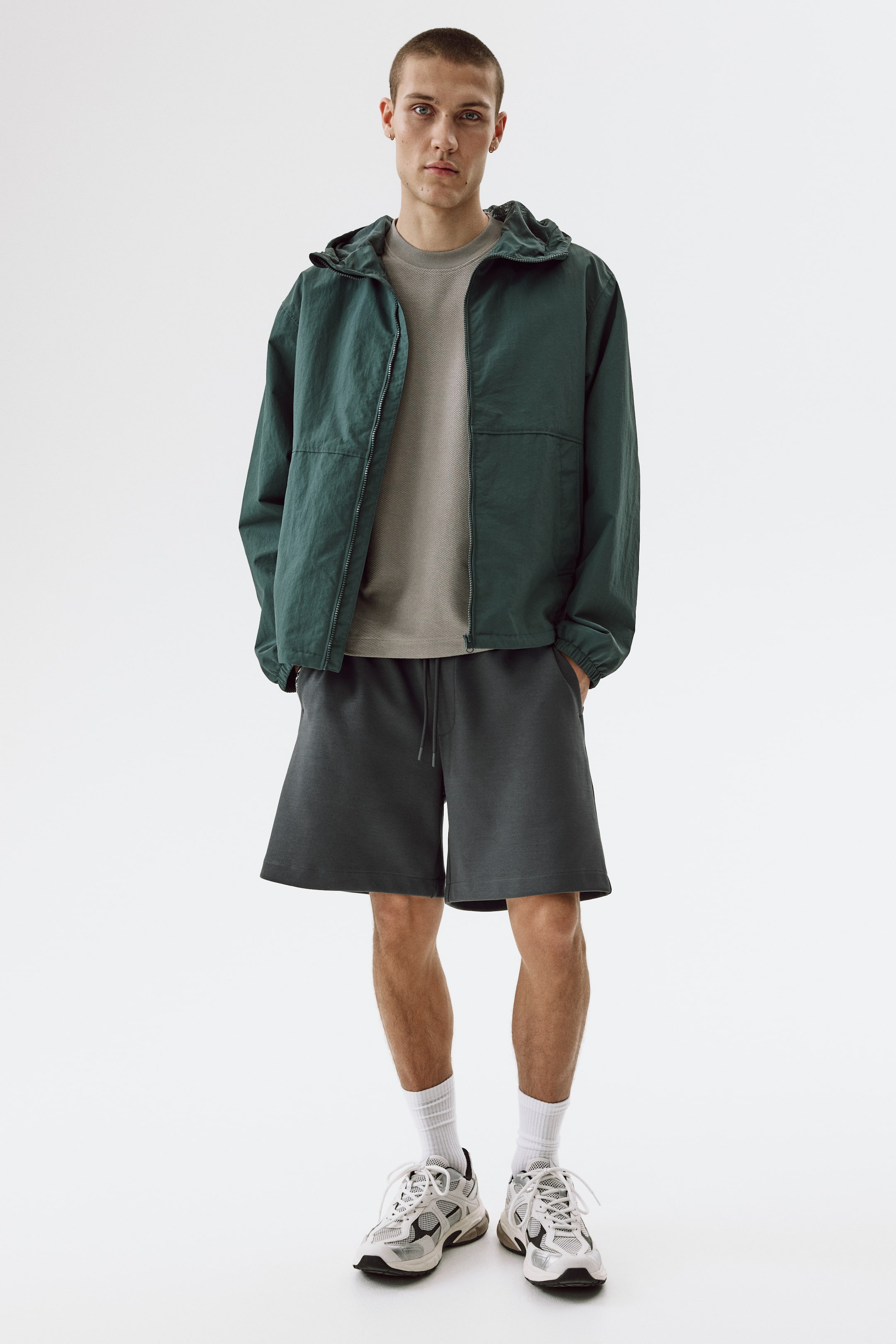 Visualizza immagine più grande: Pantaloncini grigio scuro, realizzati in un misto cotone jersey interlock e caratterizzati da un elastico in vita con coulisse, patta finta, tasche laterali diagonali e tasche posteriori a filetto, indossati dal modello. Il modello è in piedi con una mano appoggiata in una tasca laterale di questi pantaloncini a vestibilità regolare.