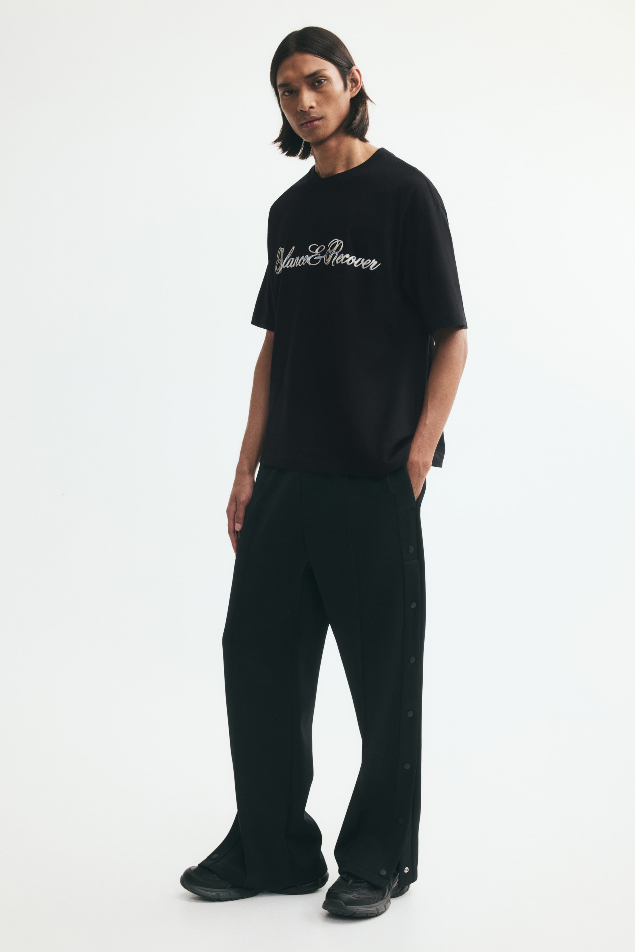 View larger image: A black relaxed-fit jersey T-shirt with a white script design printed on the chest is worn by the model. The tee features a round, rib-trimmed neckline and gently dropped shoulders, and is styled with black snap-button trousers and black sneakers.