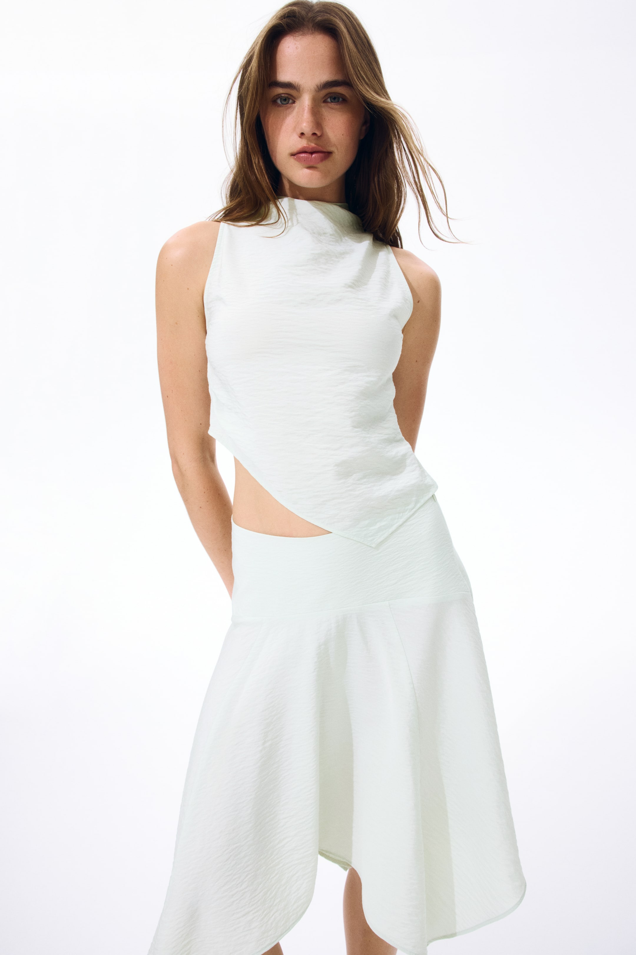 View larger image: A light green sleeveless top showcasing a cowl neckline and an asymmetric hem, with one side extending lower. It highlights a textured weave and pleats along one side, creating a gently draped effect.