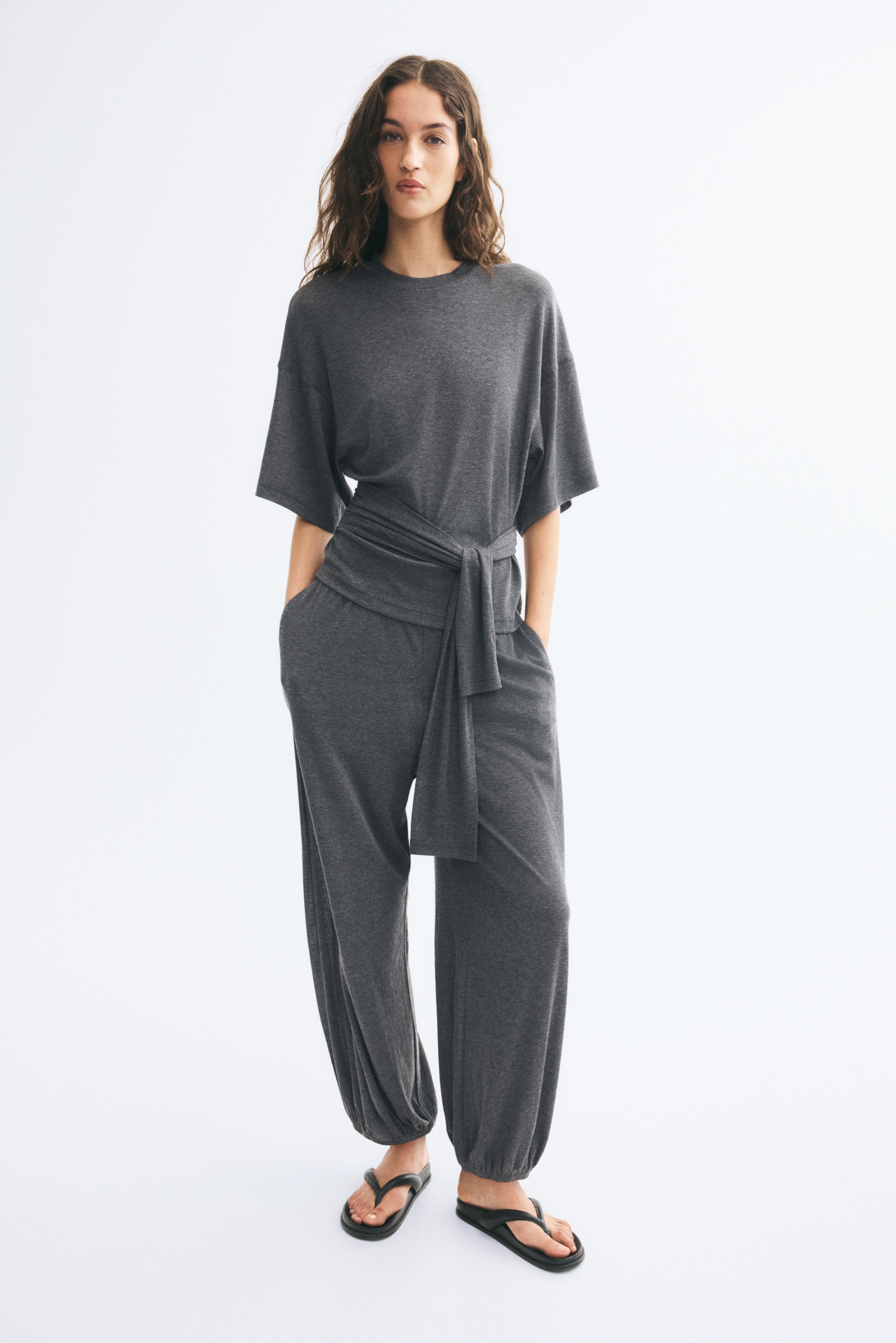 A pair of dark grey balloon-style pants is facing the front, featuring a comfortable elasticated waistband and a loose, draped fit. The soft jersey fabric gathers at the elasticated cuffs, creating a voluminous, rounded silhouette.