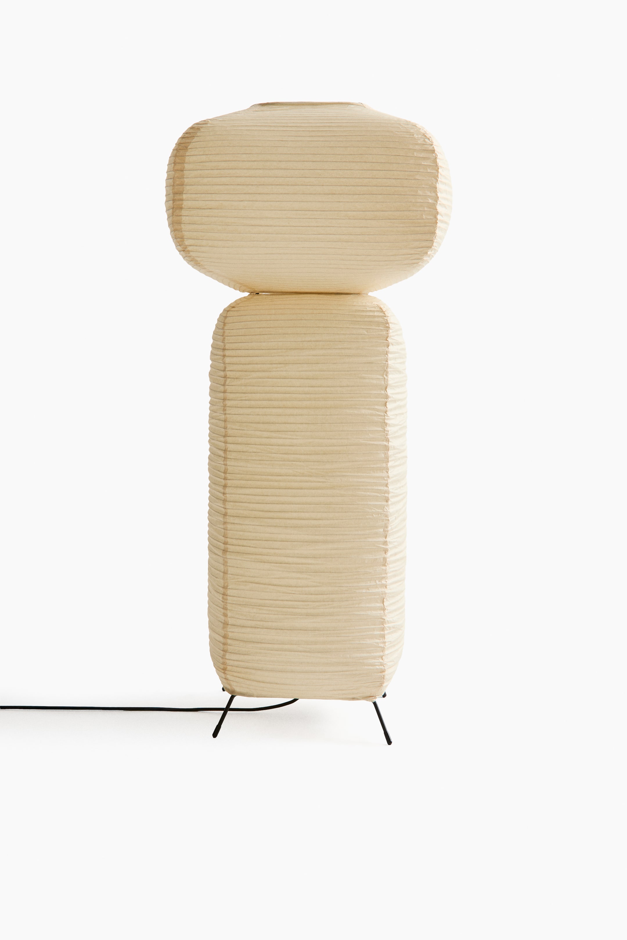 View larger image: A cream-coloured paper lamp is showcasing its front, featuring two stacked shades with horizontal pleats. The upper shade is an oval shape, resting on a taller rectangular lower shade, which is supported by a black three-legged stand with an electrical cord extending from its base.