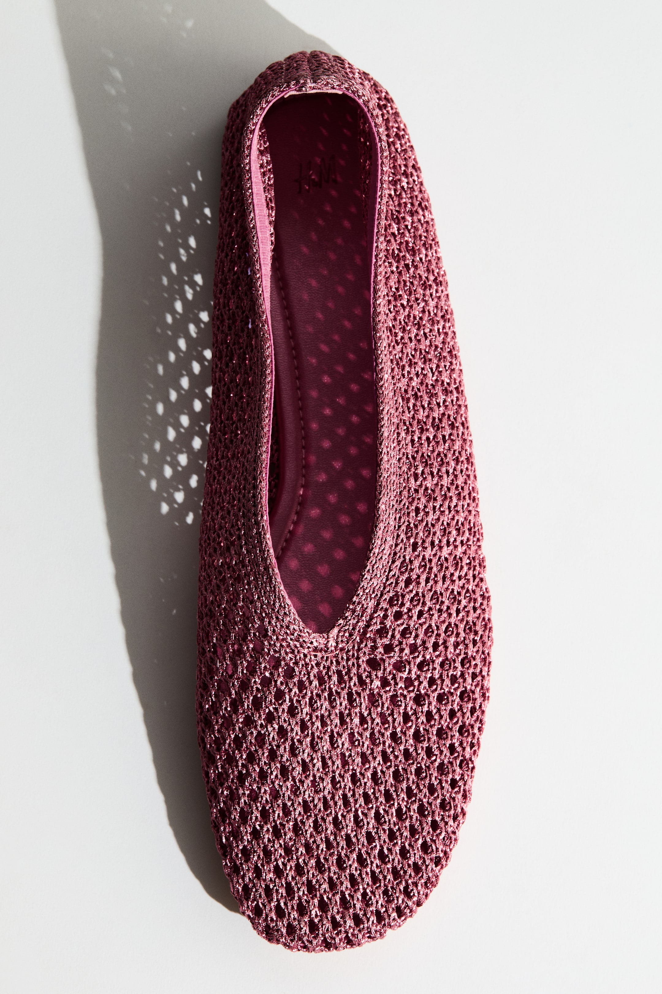 View larger image: A berry pink ballet flat showcasing an open-knit mesh upper with a pattern of small holes, a rounded toe, and a low vamp. The darker pink insole features visible perforations, with the shoe angled slightly upward and to the left.