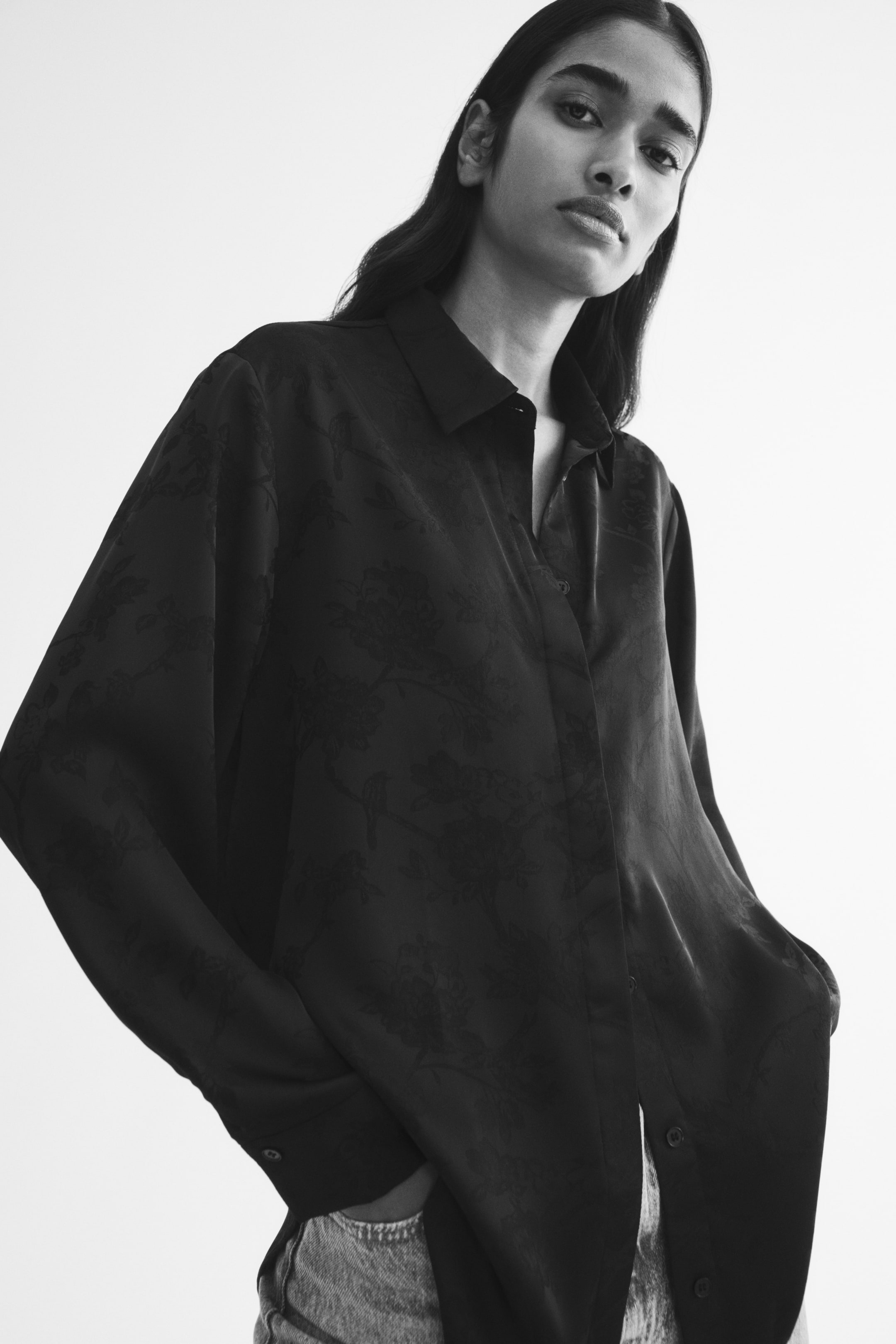 View larger image: A long-sleeved shirt in a dark jacquard weave with a subtle floral pattern is being worn by the model. The shirt features a collar, a button-down front, and long sleeves with button cuffs, and is paired with light-coloured trousers.