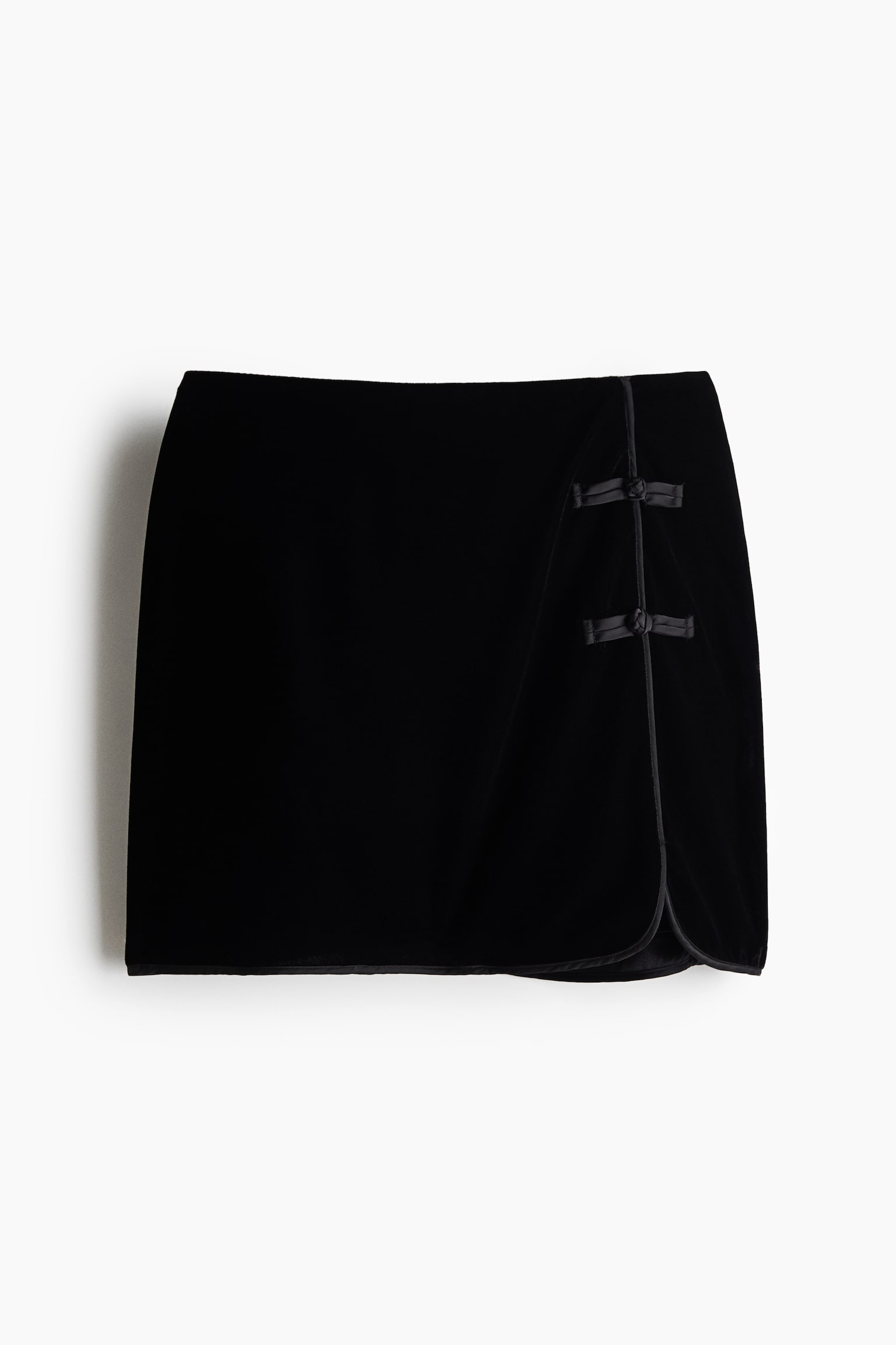 En sort mininederdel i flÃ¸jl ses forfra og fremhÃ¦ver et wrap-inspireret design med tynd satinpiping langs kanterne. Det overlappende frontpanel lukkes i venstre side med to dekorative satin-knappelukninger.