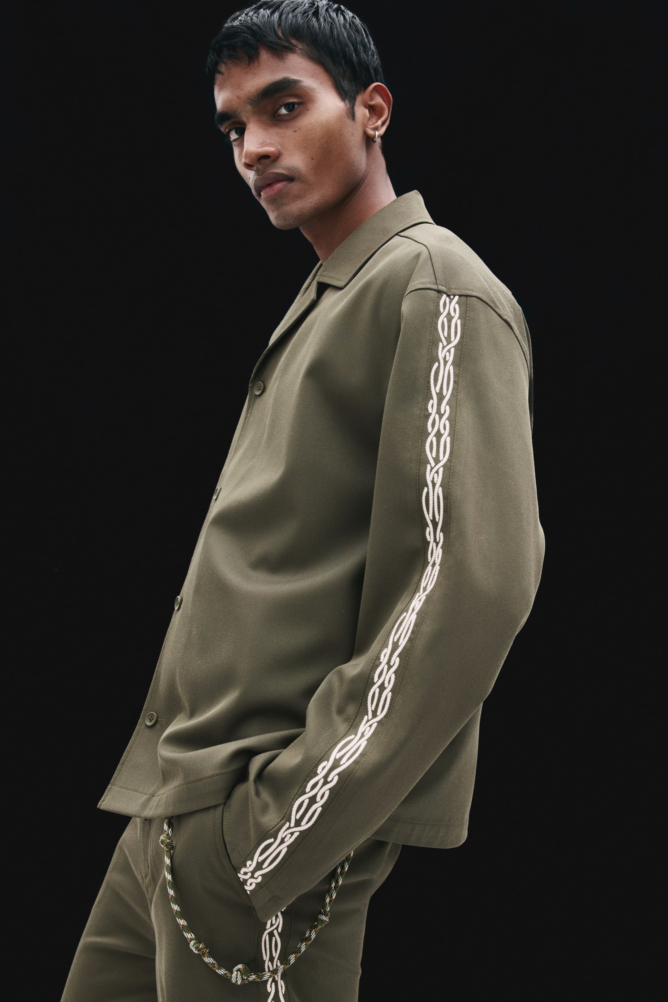 View larger image: The model is wearing an olive green overshirt featuring a resort collar, a buttoned front, and a white embroidered chain motif along the sleeve. The shirt is styled with matching trousers as the model stands in a three-quarter profile.