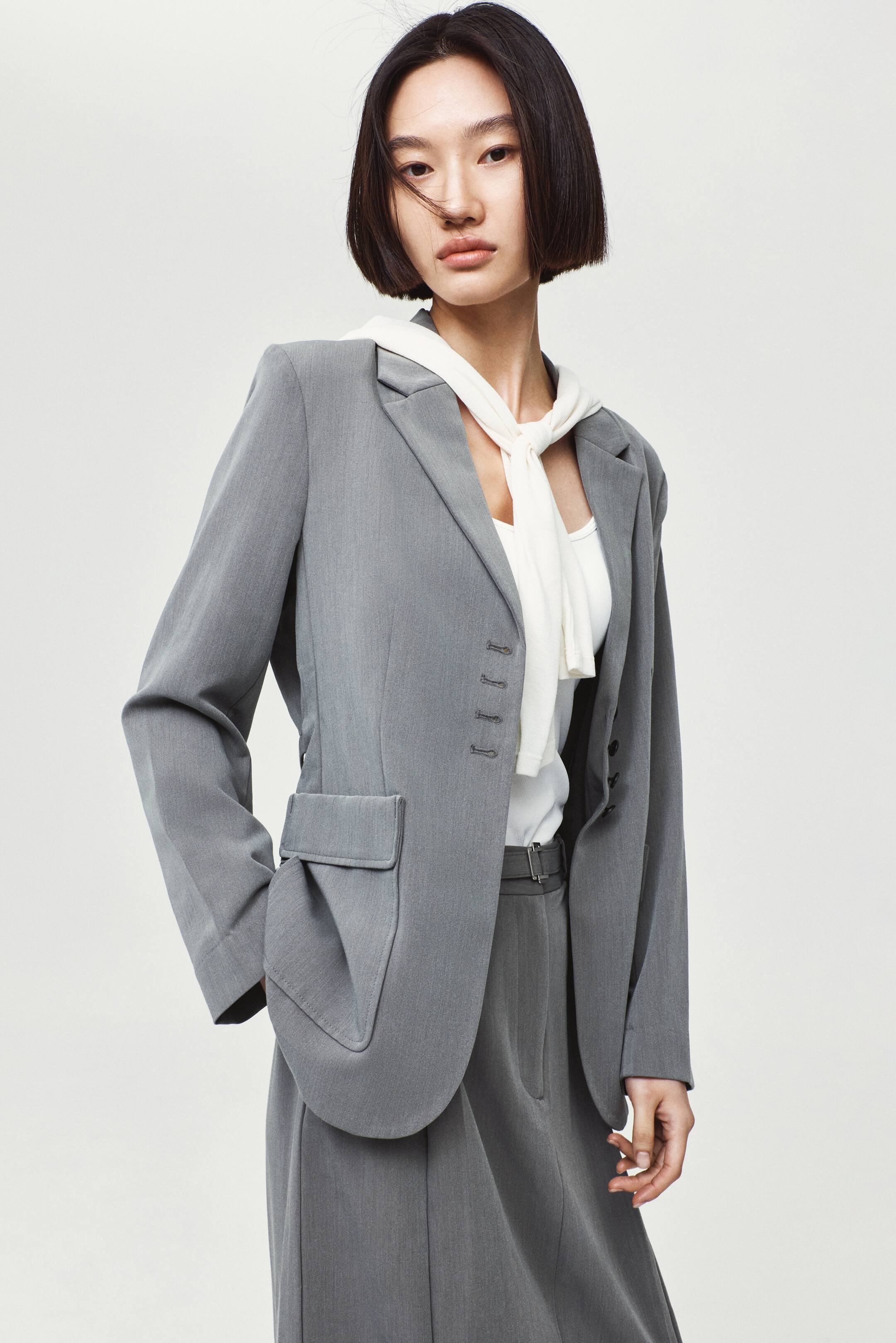 View larger image: A grey single-breasted blazer featuring notch lapels, structured shoulder pads, and front patch pockets with decorative flaps is worn by the model. The blazer is styled over a white top with a cream sweater tied around the neck, and is paired with matching grey trousers.