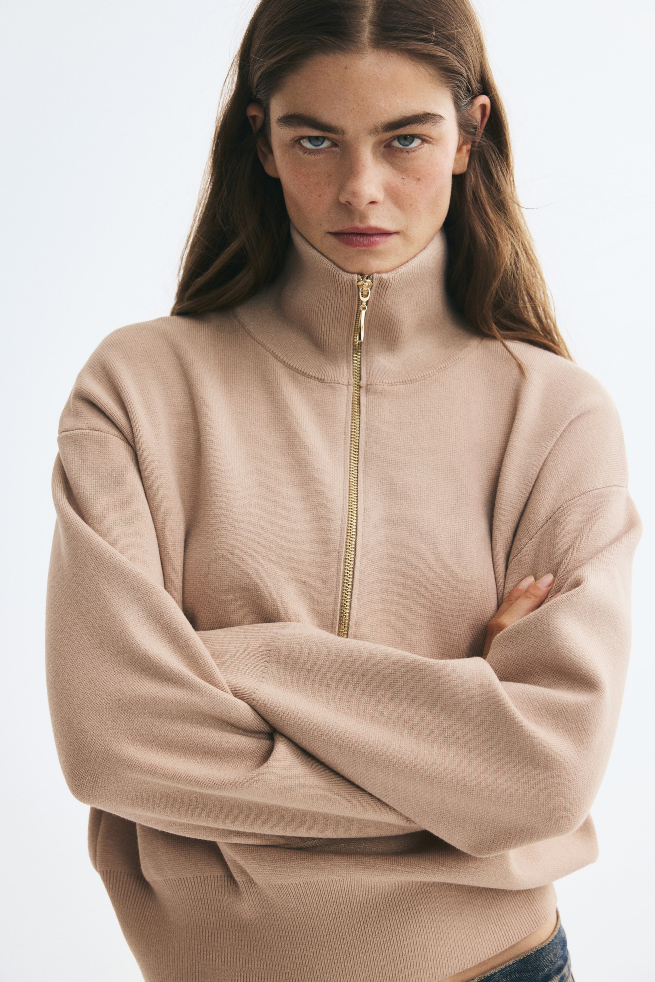 View larger image: The model is wearing a beige fine-knit sweater with a high ribbed collar and gold-toned half-zip, standing with arms crossed. The sweater features dropped shoulders, long sleeves, and a ribbed hem.