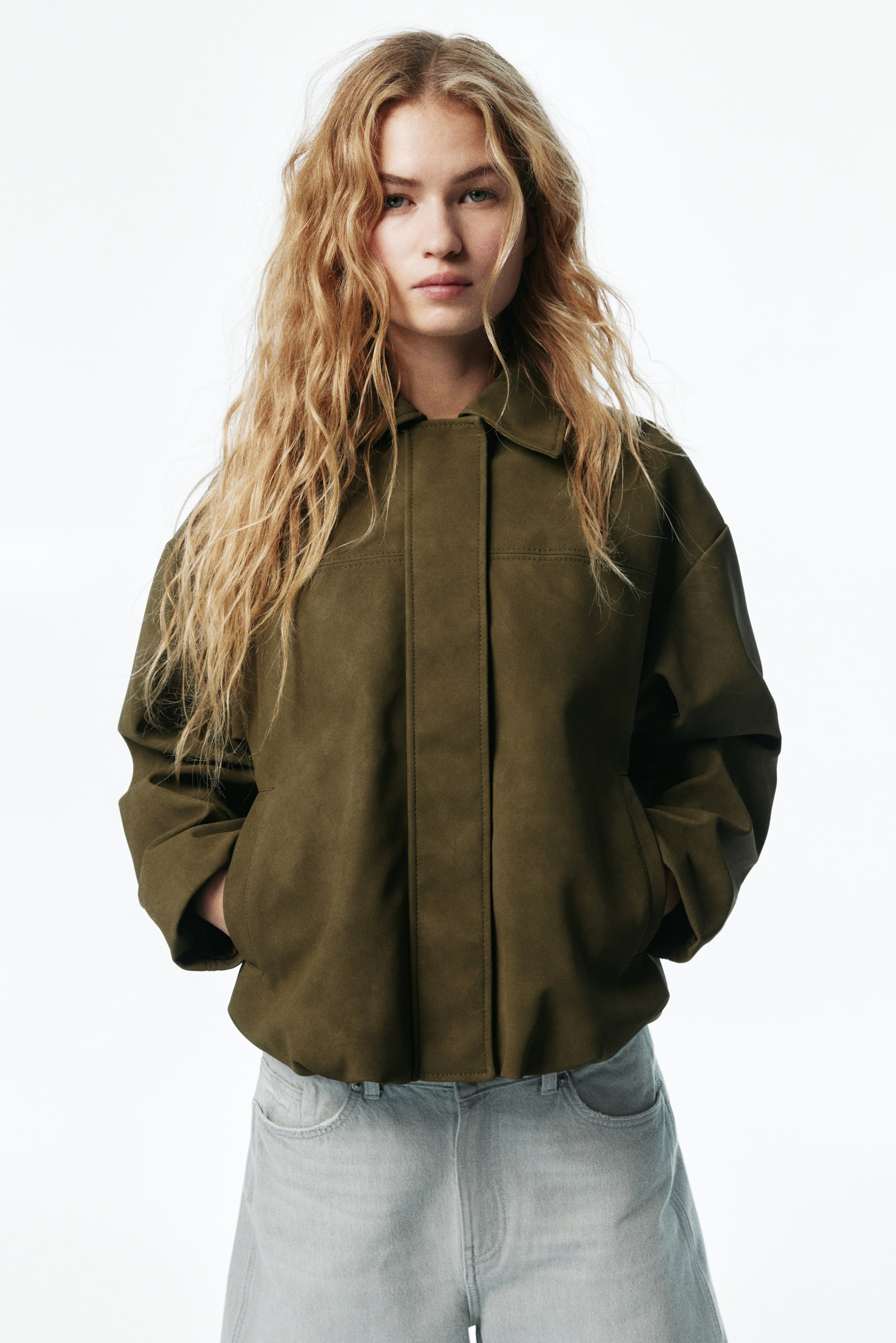 View larger image: An olive green jacket with a matte finish is featuring a pointed collar, a hidden front placket, and visible topstitching across the yoke. The jacket is presenting a relaxed fit with two side pockets and an elasticized hem, and is shown from the front.