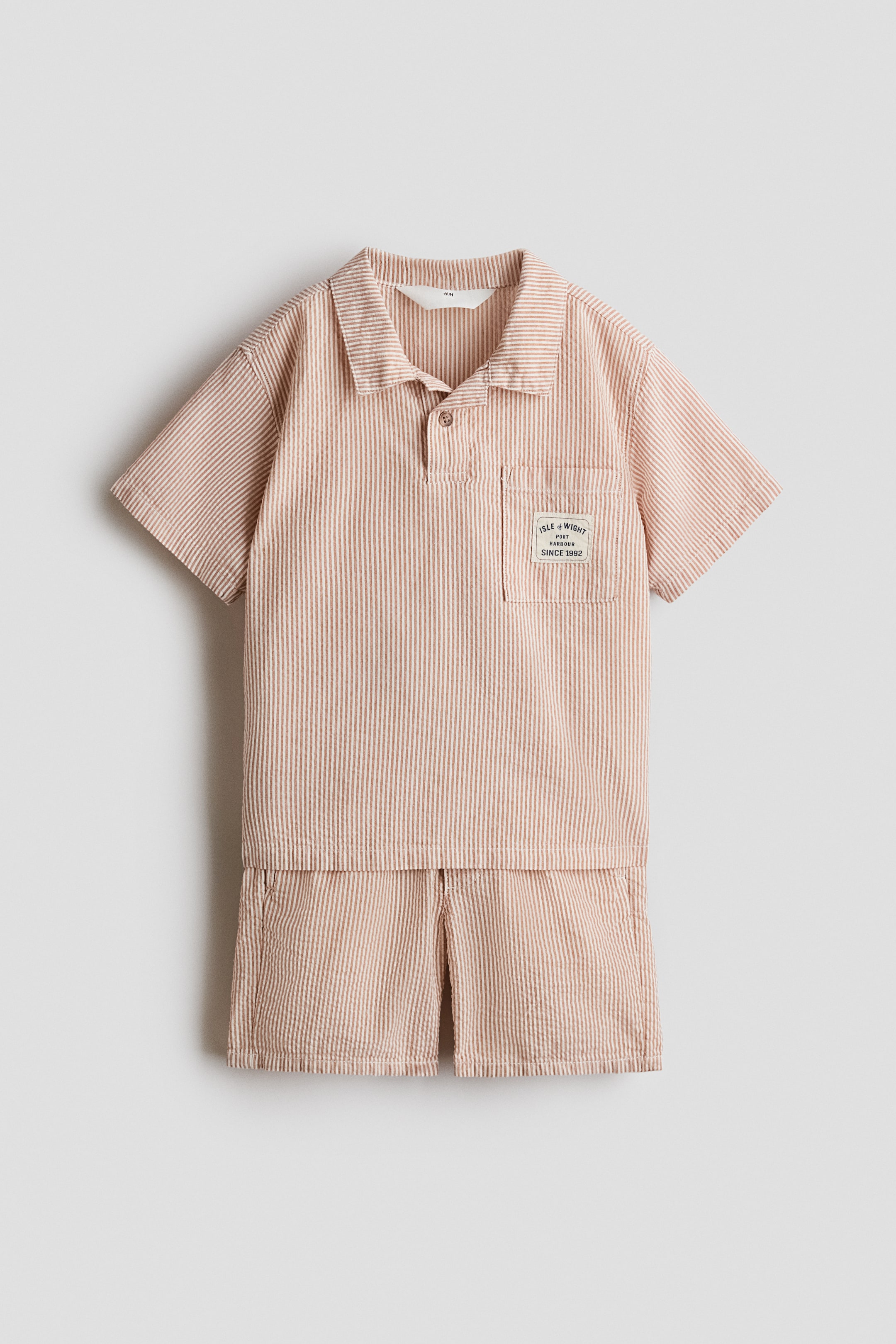 View larger image: A two-piece set comprises a short-sleeved top and matching shorts, both in light brown-and-white vertical seersucker stripes. The top is finished with a collar, a two-button placket and a patch pocket on the left chest bearing a white branded label, while the shorts offer a relaxed fit.