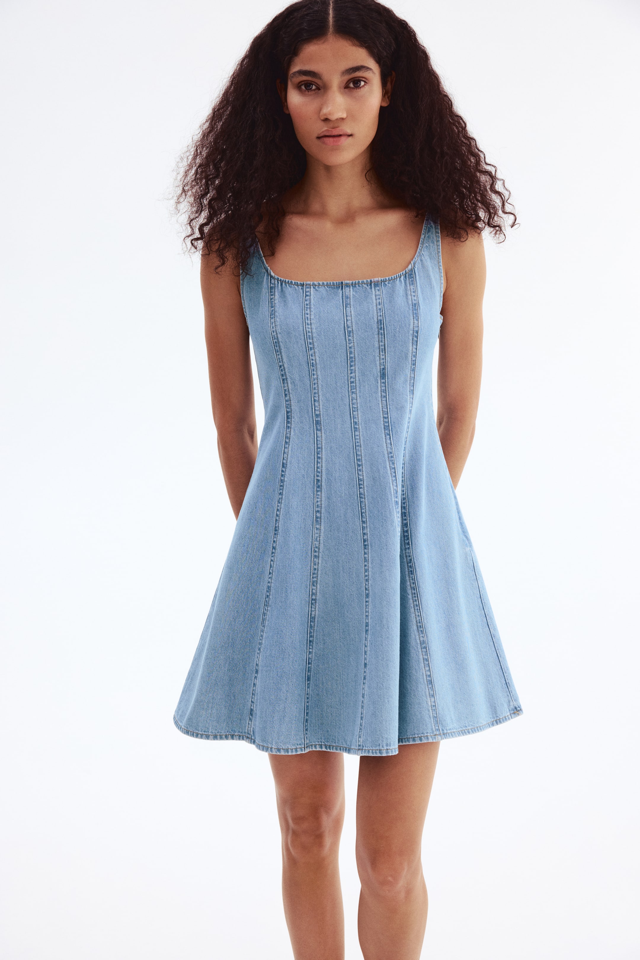 Xem ảnh lớn hơn: "Một chiếc váy mini denim xanh nhạt đang hướng về phía trước, nổi bật với cổ vuông và dây đai rộng. Nó thể hiện phần thân và chân váy ôm sát, được chia thành các mảng, và xòe ra thành dáng chữ A."