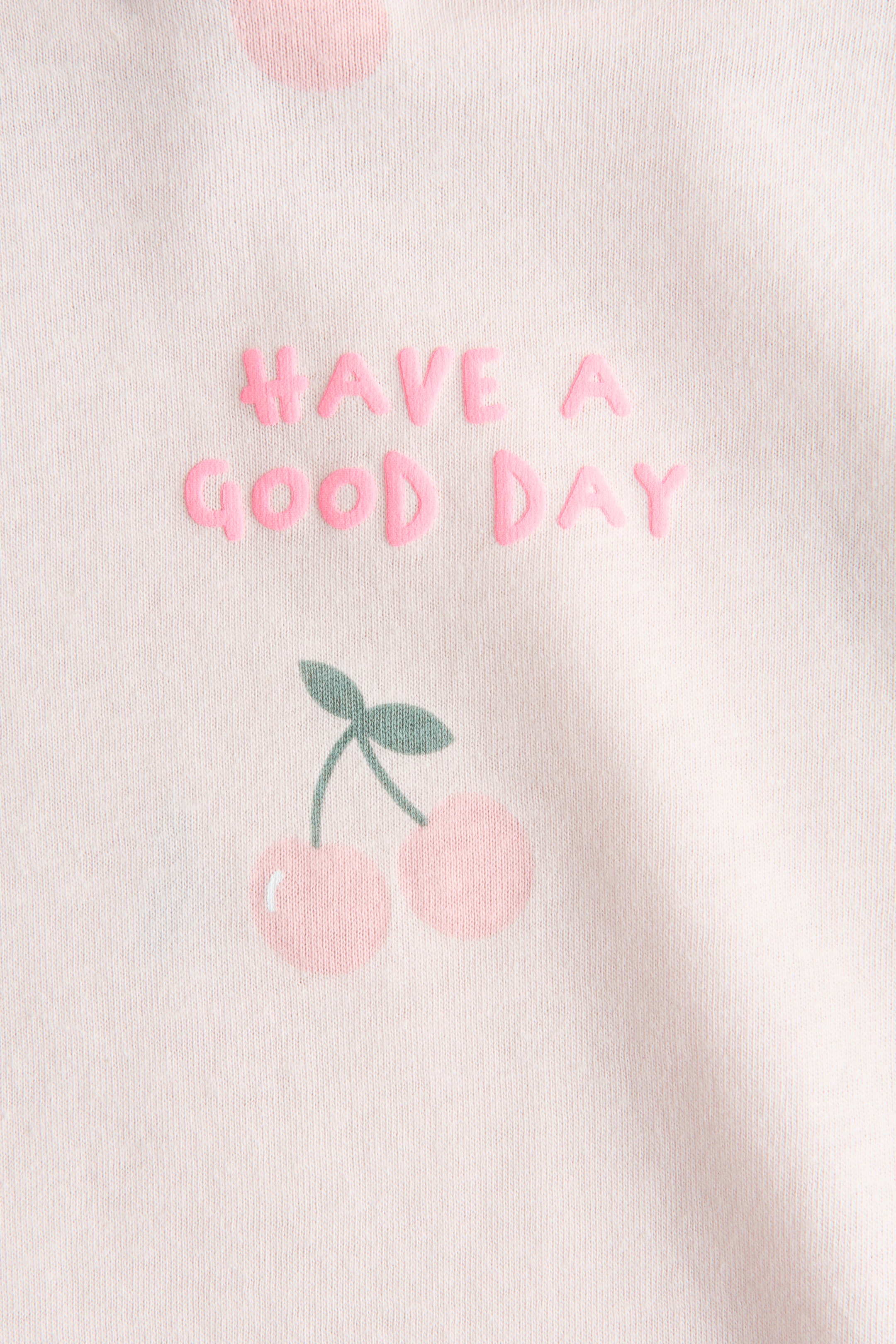 Agrandir l'image: Un tissu rose pâle affiche en relief le texte « HAVE A GOOD DAY » en rose. Sous le texte, deux cerises roses aux tiges et feuilles vertes sont visibles, ainsi que de grands pois roses pâles et discrets.