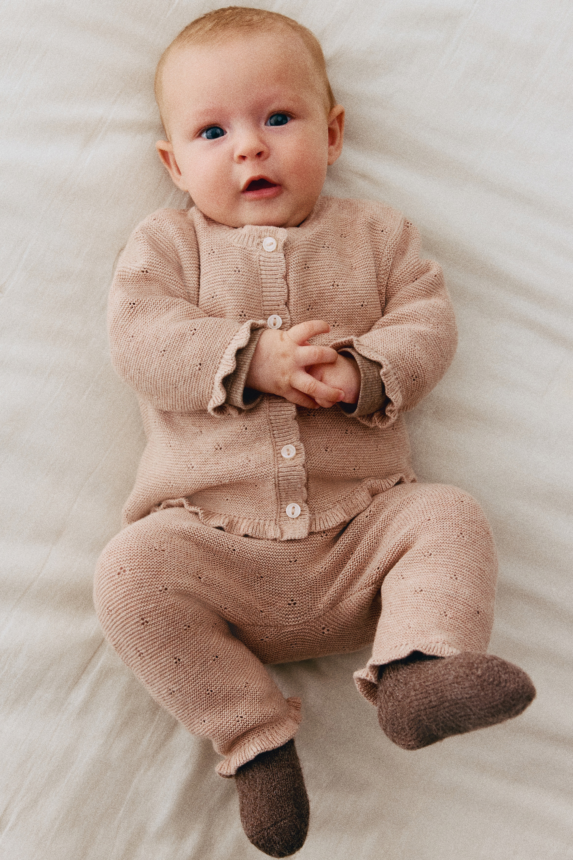 A two-piece knit baby set in dusty pink is displayed front-facing, featuring a cardigan and matching pants with a pointelle knit pattern. The long-sleeved cardigan has a button-front closure with ruffled edges at the collar, cuffs, and hem, while the matching pants feature ruffled ankle cuffs.
