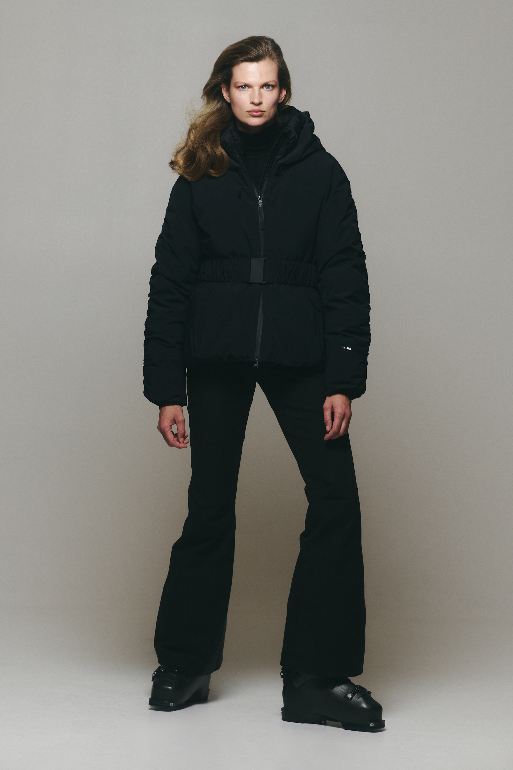 View larger image: A black hooded ski jacket with a front zipper and an integrated elastic belt with a center buckle is being worn by the model. The jacket is styled with matching black flared ski pants and black ski boots.