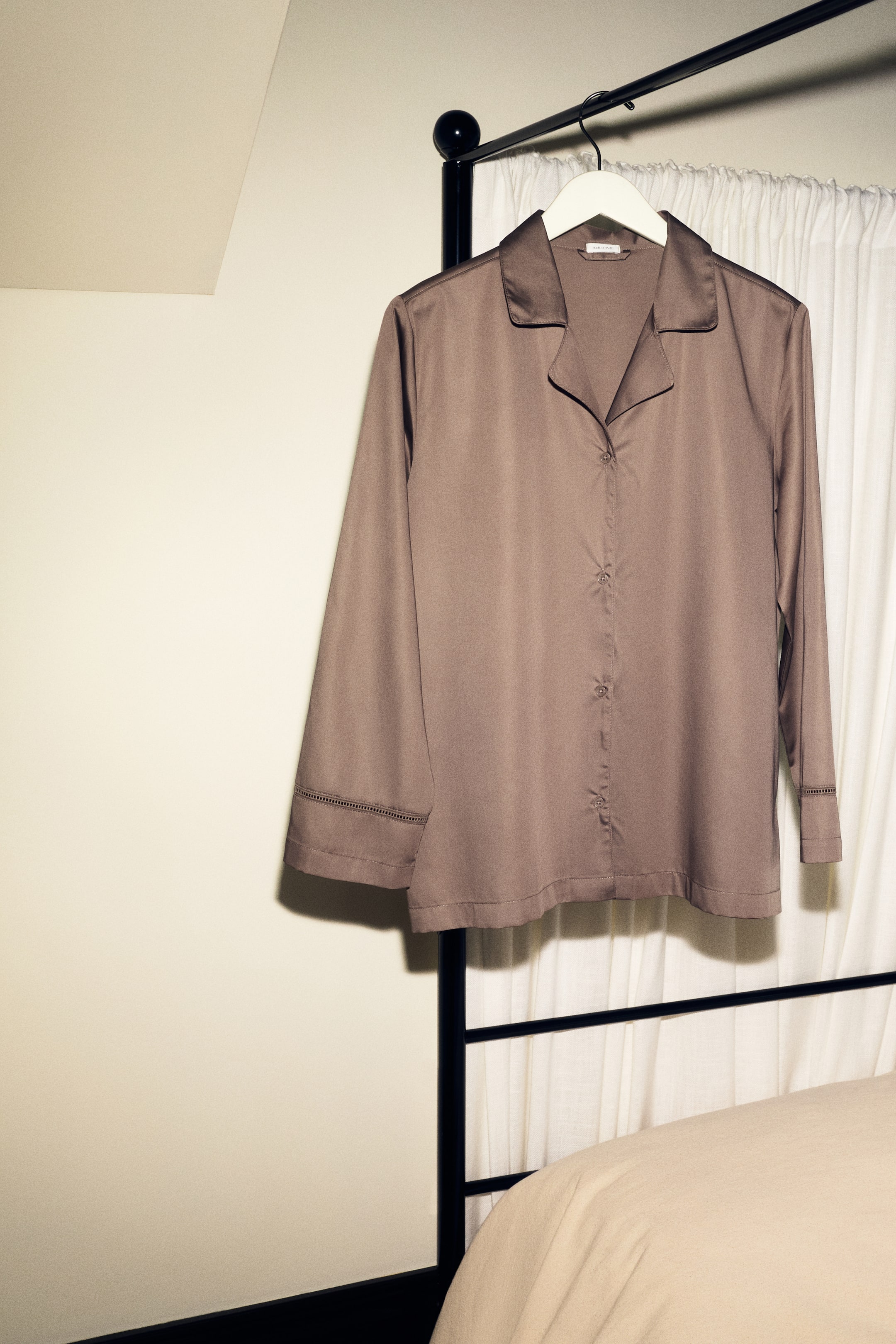 View larger image: A long-sleeve pajama shirt in taupe sateen is hanging front-facing, showcasing its subtle sheen. The shirt features a resort collar, a button-down front, and delicate ladder lace insets detailed near the cuffs.