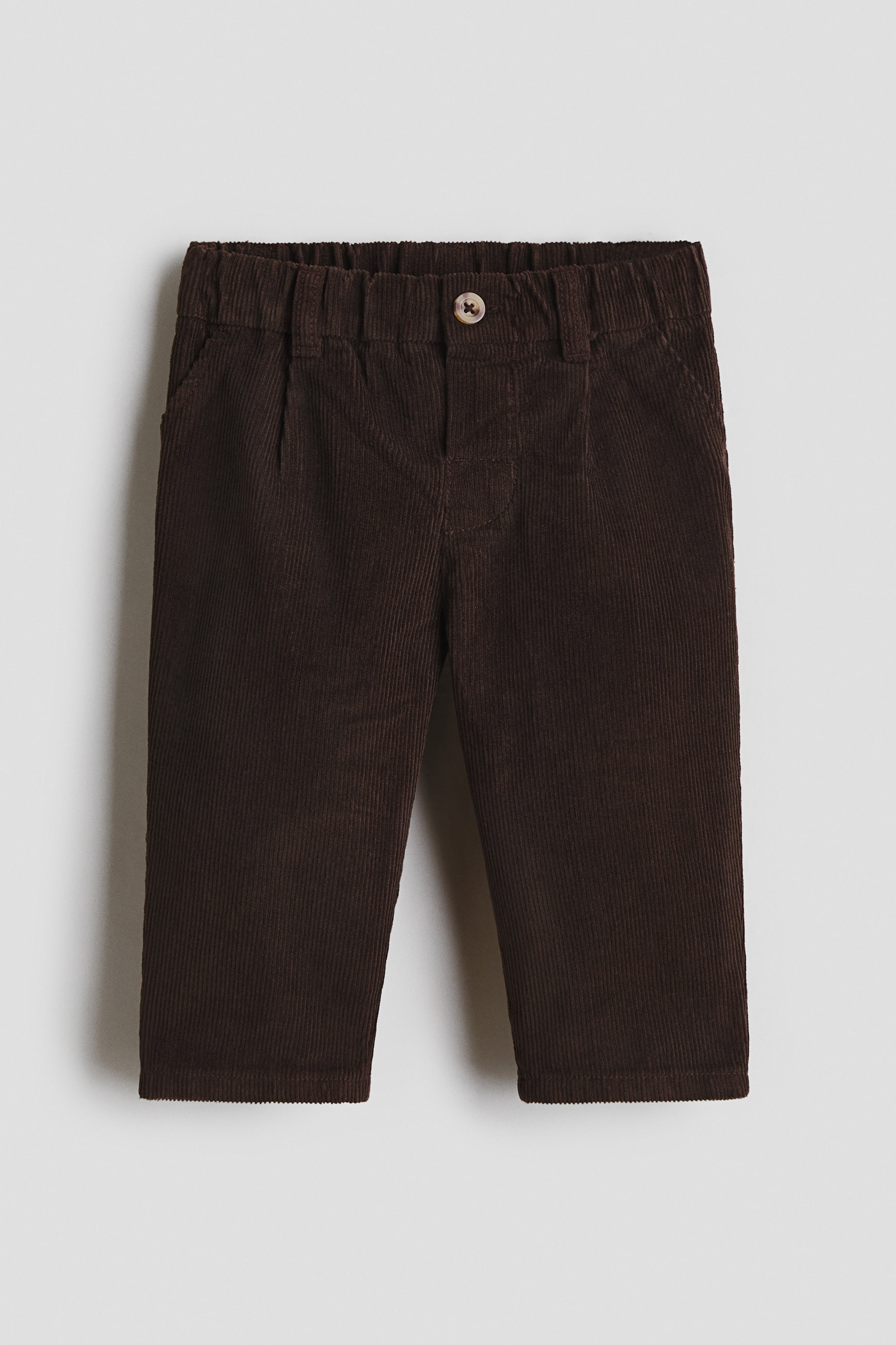 Ver imagen más grande: Pantalones de pana marrón mostrados en vista frontal, con cintura elástica y cierre de un solo botón. Se aprecian dos bolsillos delanteros inclinados, y unos pliegues verticales se extienden por debajo de la cintura.