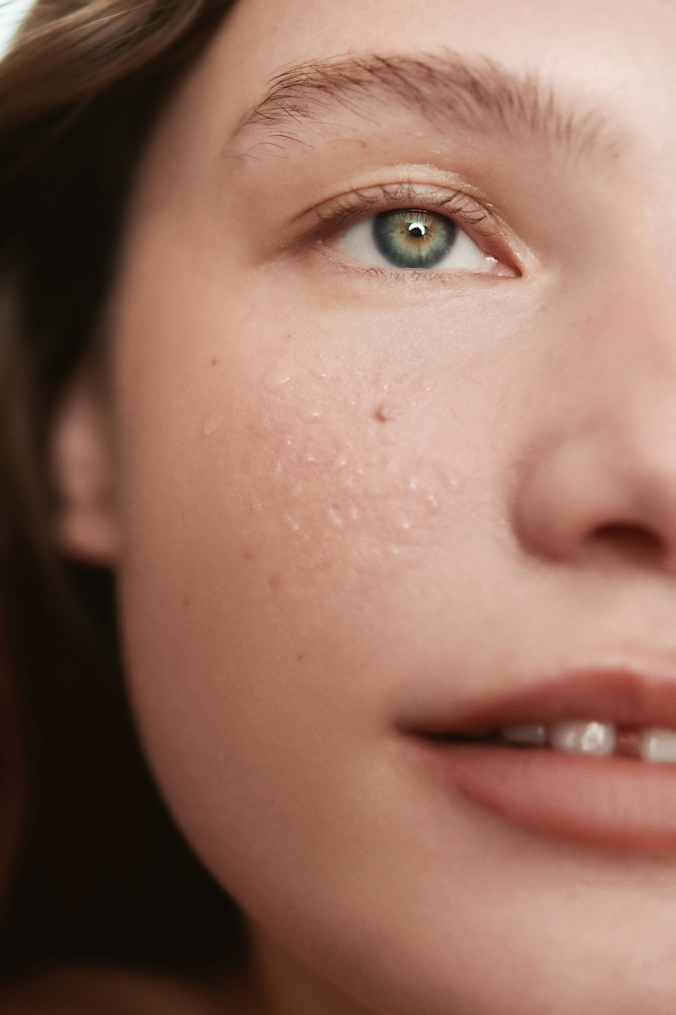 View larger image: A close-up of a person's face shows the right side, highlighting a green-hazel eye with visible water droplets on the cheek. The skin appears smooth with some texture and pores, and part of the lips and teeth are visible.