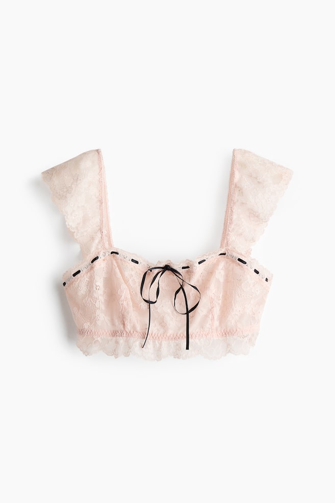 A pink lace bralette is shown from the front, featuring wide ruffled lace straps and a black ribbon laced through small loops along the top edge of the front panel, tied into a bow at the centre. A scalloped lace trim finishes the bottom hem of the bralette. - 1