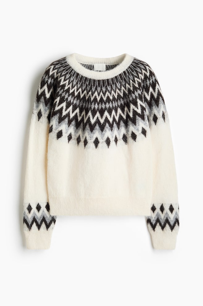 En kremfarget, myk strikkegenser vises forfra, med bÃ¦restykke i Fair Isle-stil i svart, grÃ¥tt og hvitt med chevron- og diamantmÃ¸nstre rundt halsutringningen og skuldrene. De lange ermene har matchende mÃ¸nstrede felt over de ribbestrikkede mansjettene, som komplementerer den ribbestrikkede rundhalsen og kanten. - 1