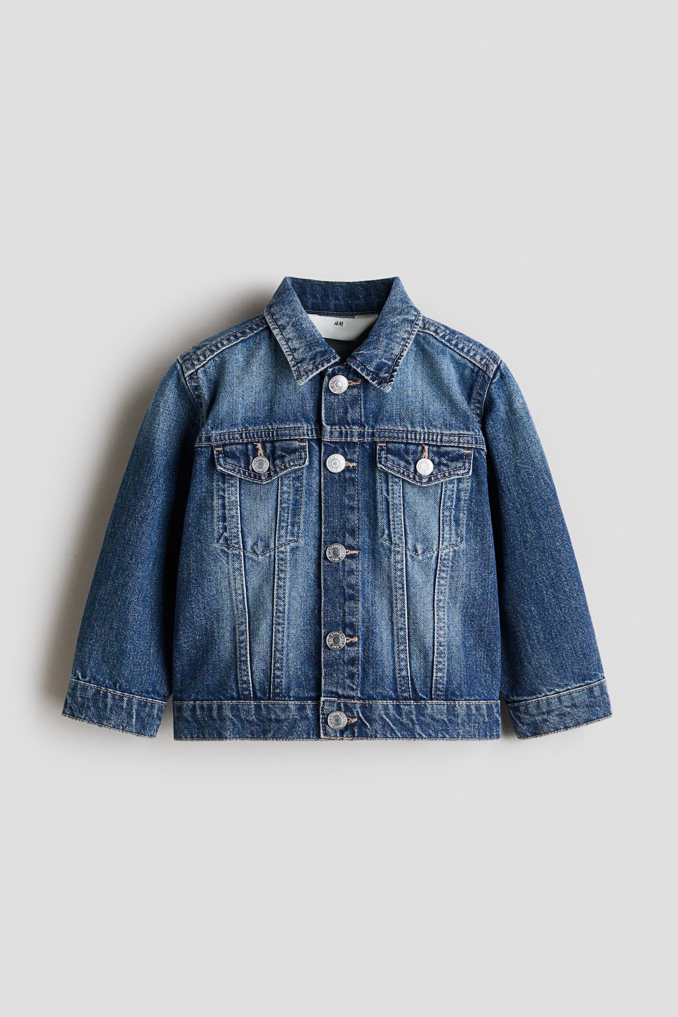 View larger image: The classic denim jacket is shown facing front, crafted from blue cotton denim with a faded wash. It features a turn-down collar, press-studs running down the front and on the cuffs. Two decorative chest pockets are visible, each with a flap secured by a press-stud.