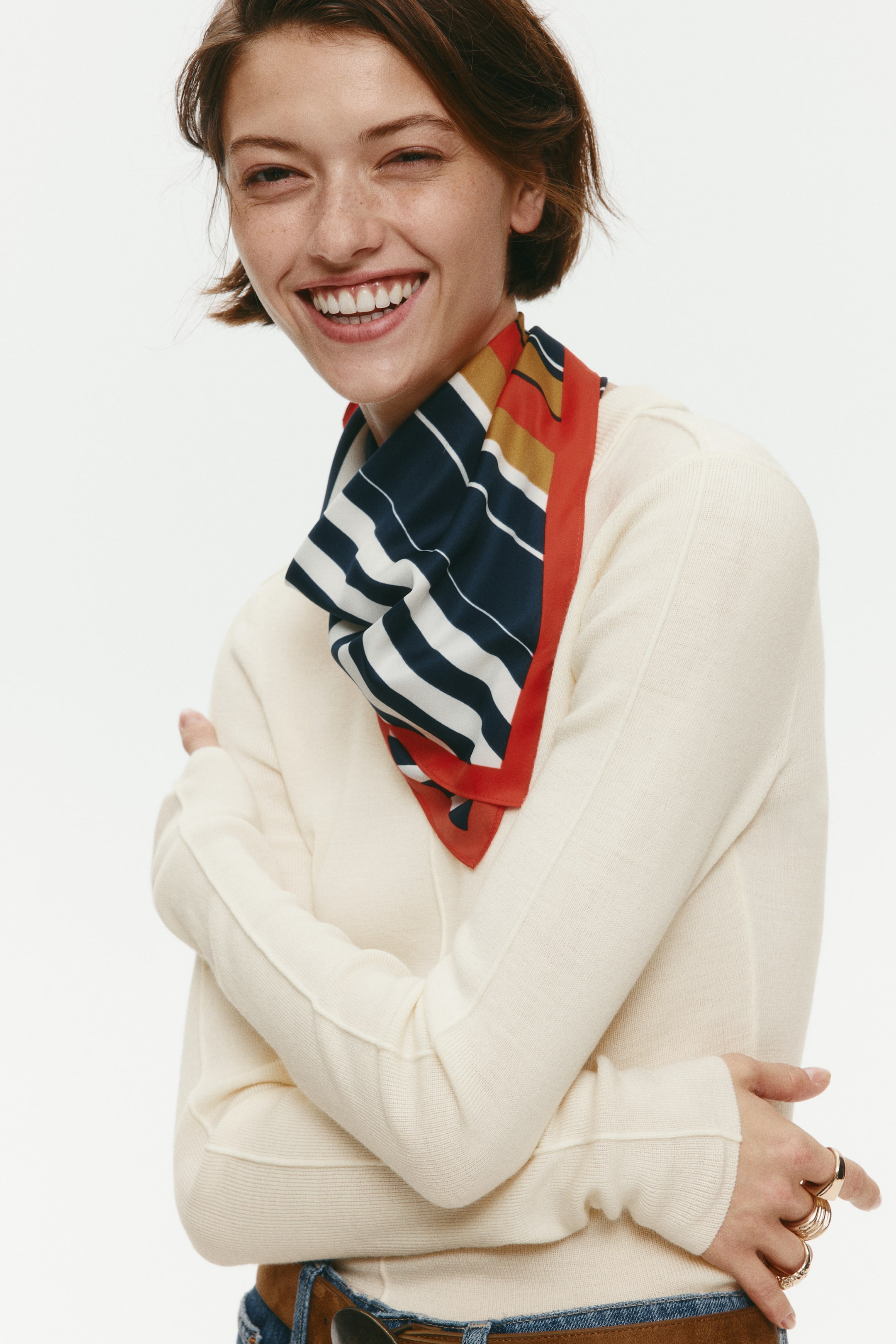 View larger image: A cream-colored knit top featuring long sleeves and visible linked seams is being worn by the model, who is crossing their arms. The top is styled with a striped neck scarf in navy, white, red, and mustard yellow.