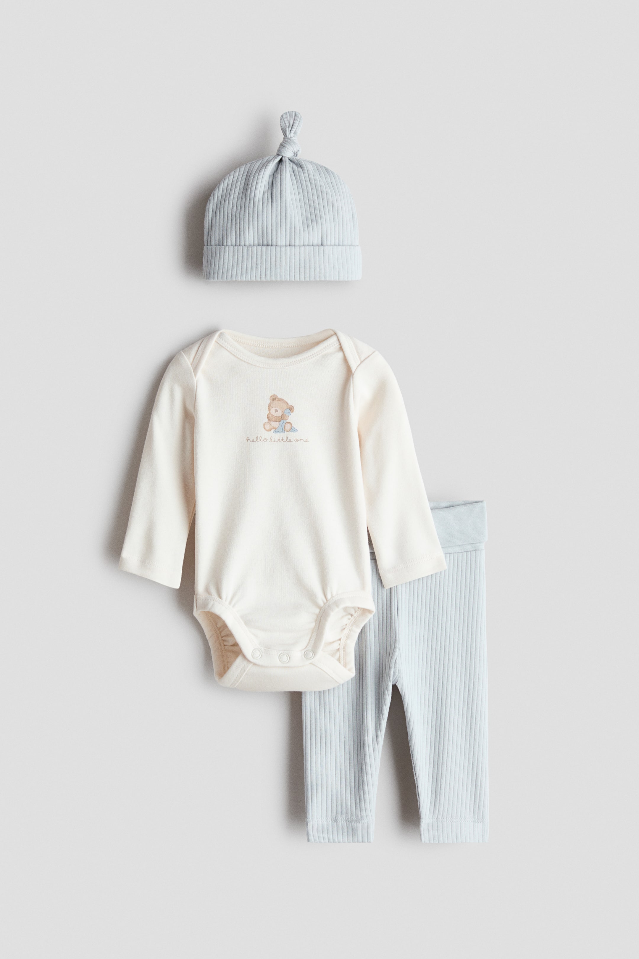 View larger image: A three-piece baby clothing set featuring a cream long-sleeved bodysuit with a round neckline, a teddy bear graphic, and "hello little one" text on the front, along with press-studs at the gathered leg openings. It is paired with light blue ribbed trousers that have a foldover waistband and cuffed hems, as well as a matching light blue ribbed hat with a knotted top.