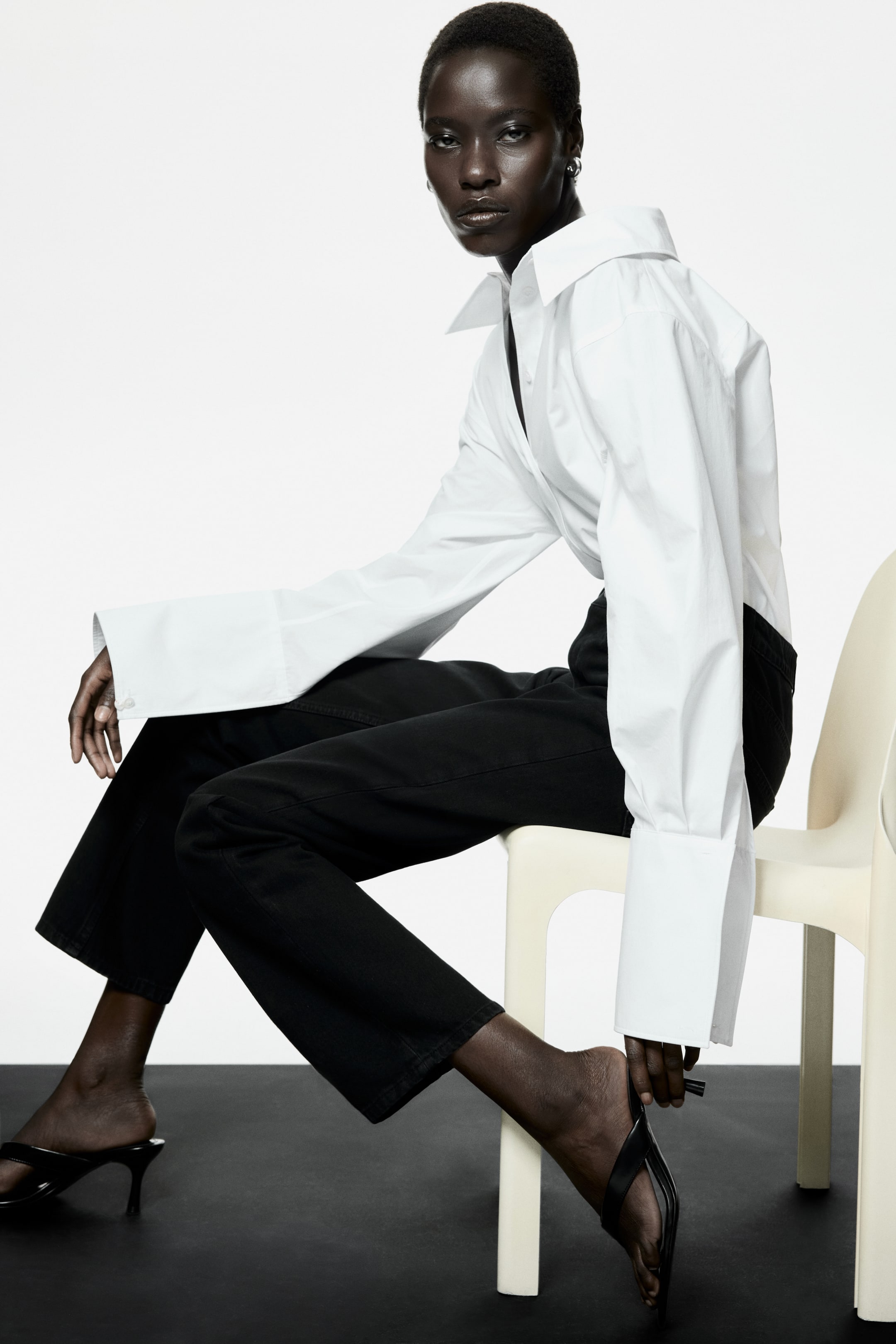 Agrandir l'image: Une chemise blanche oversize à boutons est dotée d'un col étalé montant et de larges poignets profonds. Un pantalon noir raccourci présente une coupe droite et décontractée, et des sandales noires à talons sont portées.