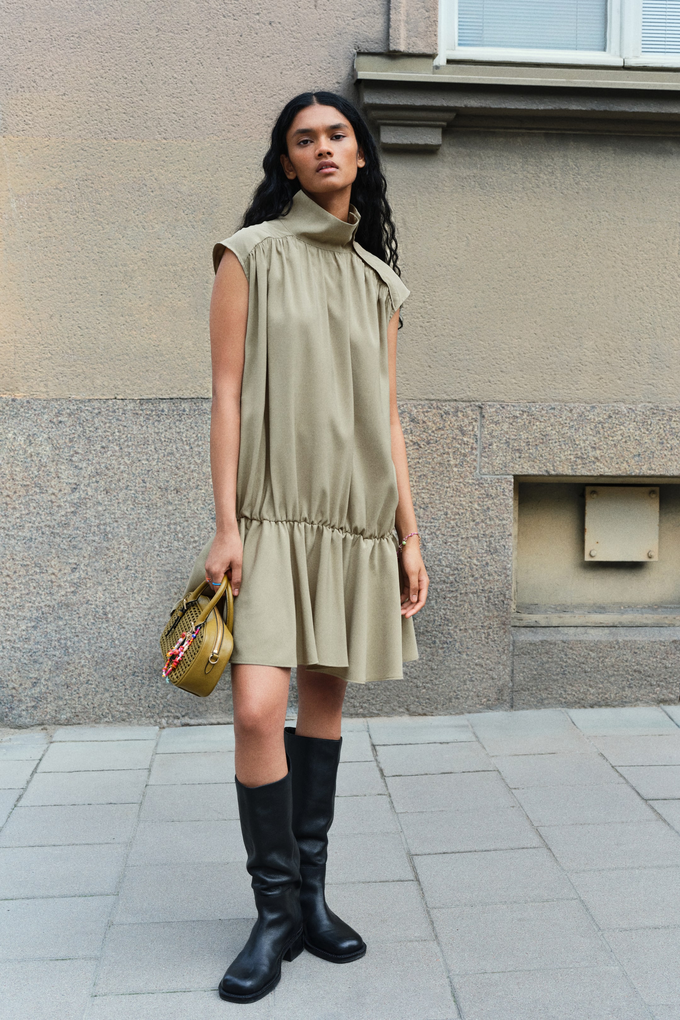 Größeres Bild ansehen: Das Model trägt ein kurzes, ärmelloses olivgrünes Kleid mit hohem Stehkragen, lockerer Passform und einer tief sitzenden, elastischen Taille mit ausgestelltem Rock. Das Kleid wird mit schwarzen, kniehohen Stiefeln kombiniert, und das Model trägt dazu eine kleine, passende olivgrüne Handtasche.