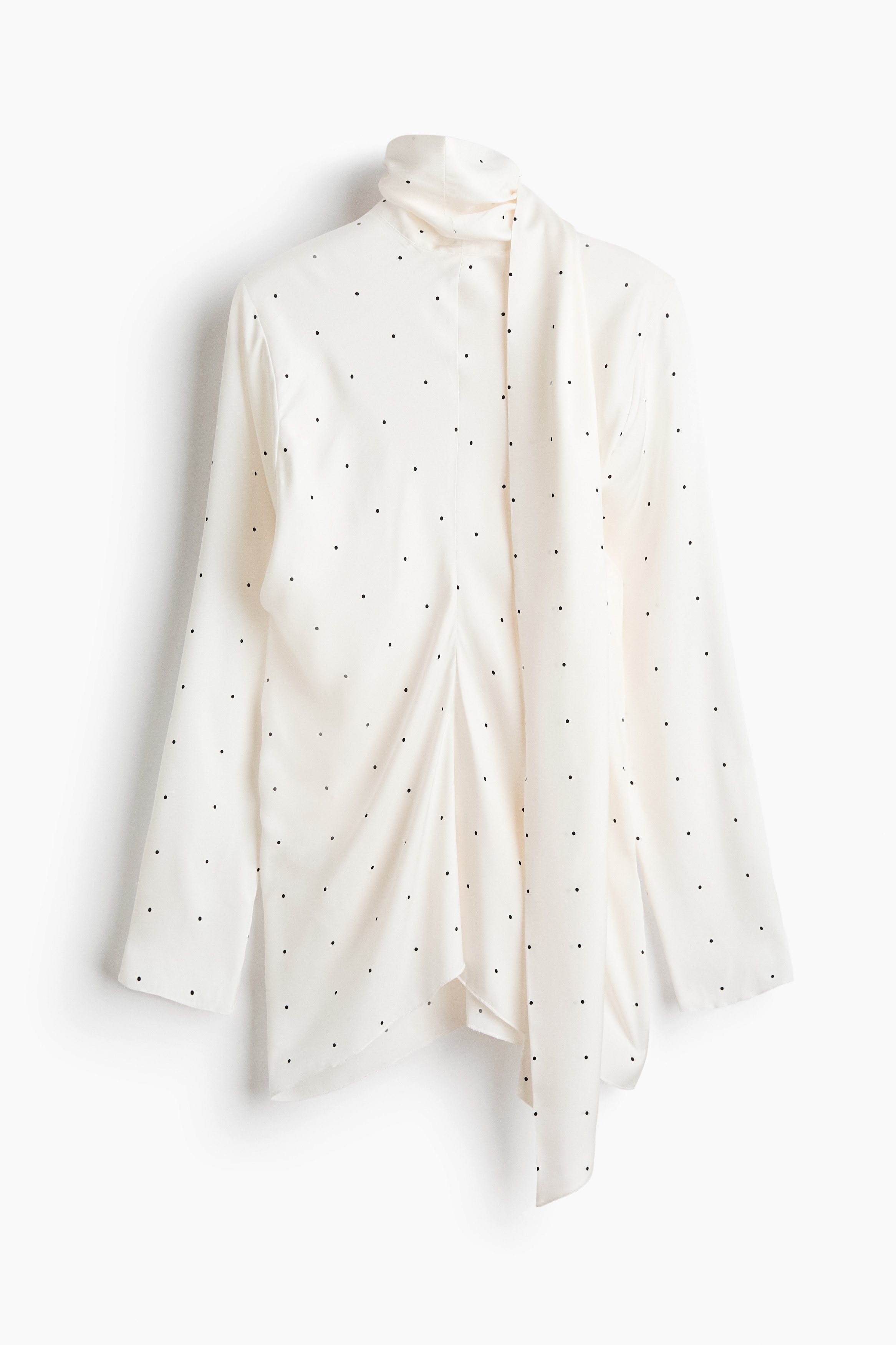 The model is wearing a white polka-dot blouse, featuring a high scarf collar with a long draped tie hanging down the front. The blouse has long sleeves and a gathered centre seam, and is styled with black jeans.