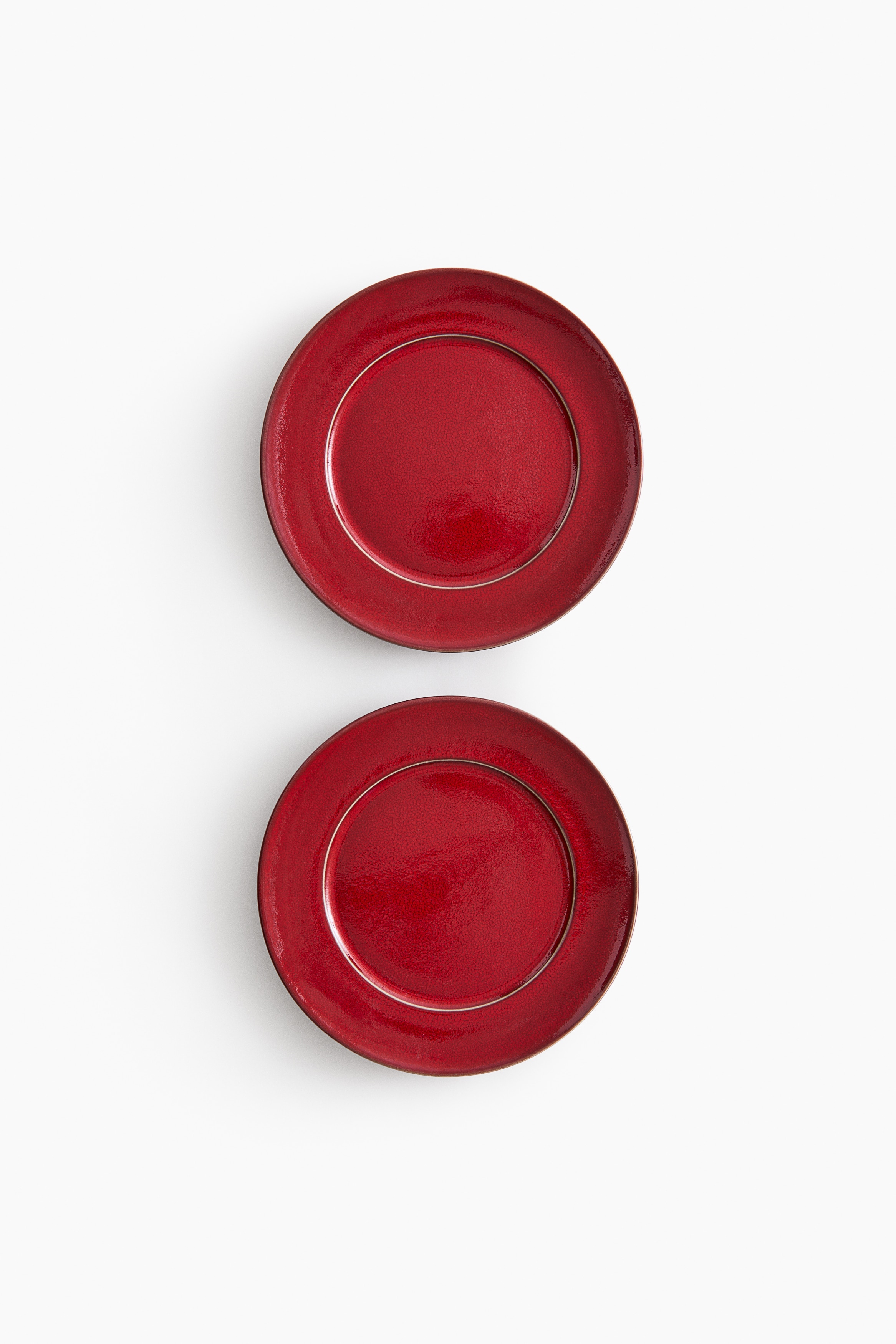 Red ceramic plates are shown from an angled top-down view, featuring a vibrant red glossy finish with a distinctive crackle glaze texture. The plates have a raised rim and a recessed centre well, with a thin, lighter-toned edge highlighting their circular shape.