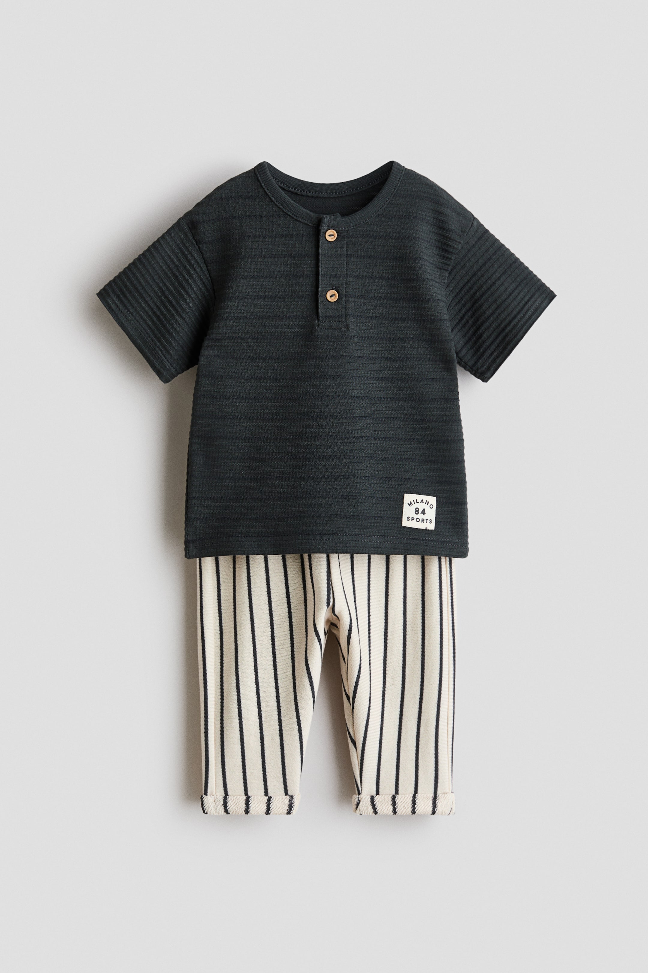 View larger image: The product is a set featuring a dark grey, short-sleeved Henley shirt, crafted from textured jersey fabric, showcasing a round trimmed neckline and a two-button placket at the front. It is paired with cream trousers, featuring black vertical stripes and sewn-in turn-ups at the hems.