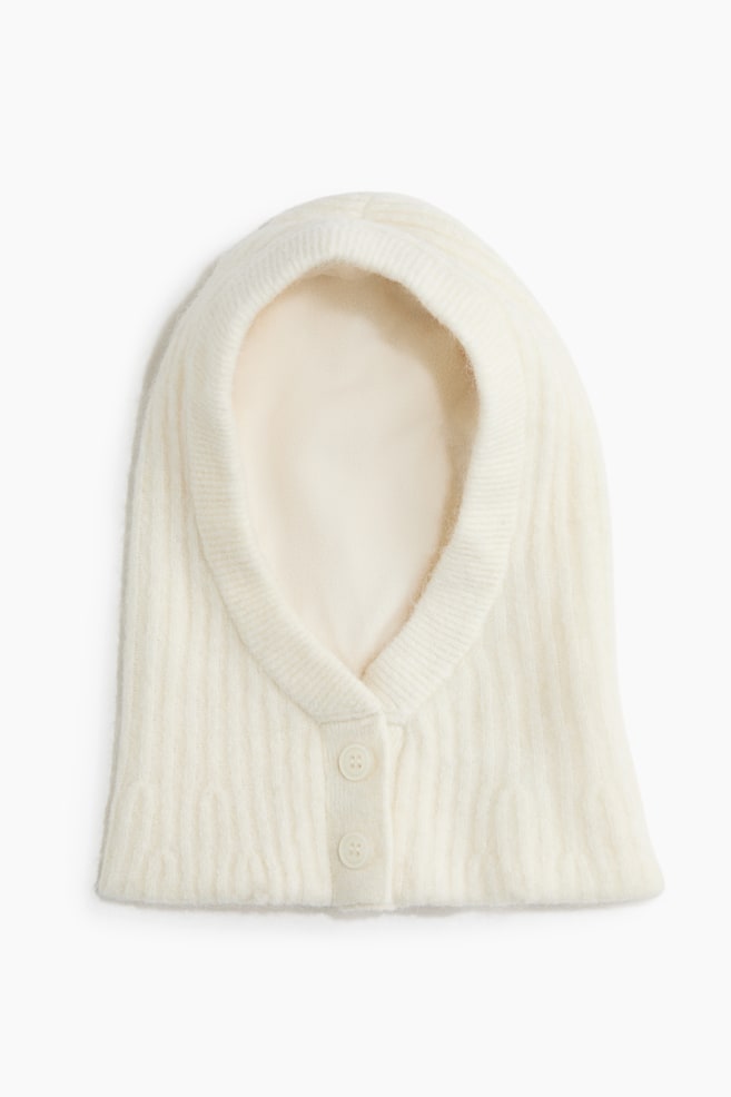Une capuche en maille de couleur crÃ¨me est posÃ©e Ã  plat, orientÃ©e vers lâavant, mettant en valeur une texture en tricot cÃ´telÃ© et une doublure intÃ©rieure douce. La capuche prÃ©sente une bordure cÃ´telÃ©e autour de lâouverture du visage ainsi quâune courte patte de boutonnage Ã  lâavant avec deux boutons assortis de couleur crÃ¨me. - 1