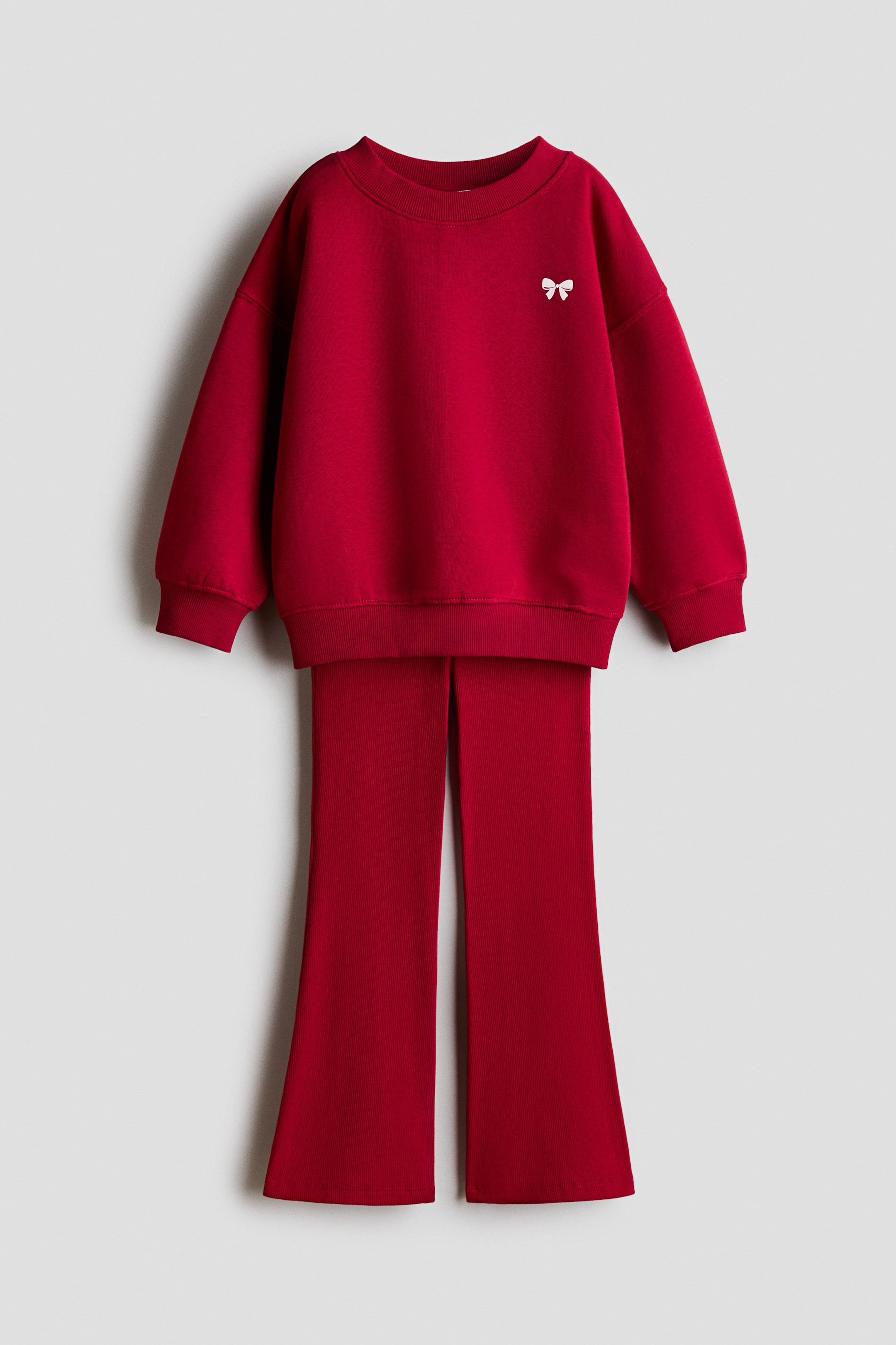 View larger image: A red two-piece set is shown in a front-facing view, featuring a crewneck sweatshirt and matching ribbed leggings. The sweatshirt has dropped shoulders, ribbed cuffs, a ribbed hem, and a small white bow on the left chest, while the leggings flare out at the hems.