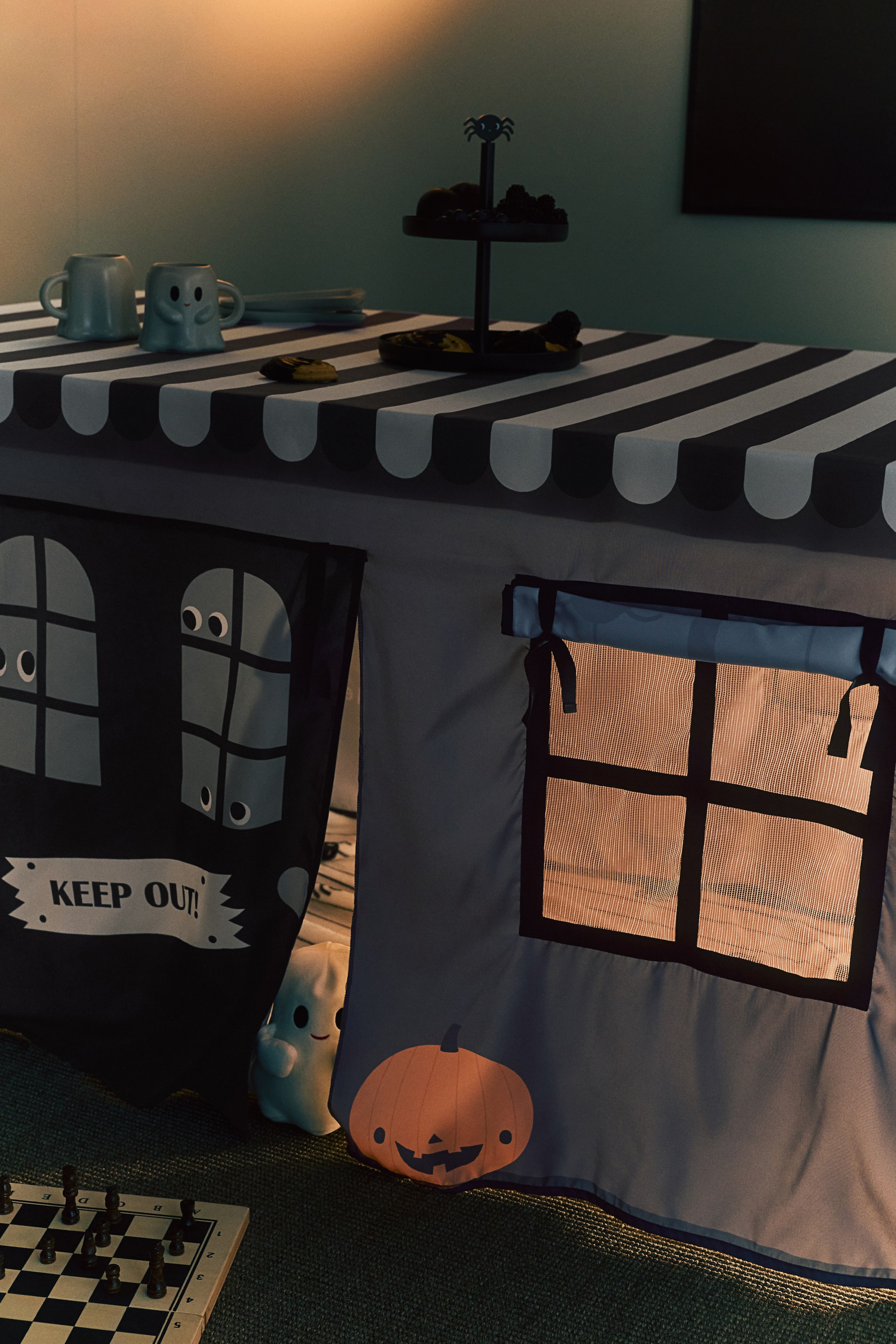 View larger image: A woven table tent in a Halloween theme is draped over a table, featuring a black-and-white striped roof with a scalloped edge. The grey front of the tent showcases a mesh window with a rolled-up fabric curtain, a printed orange pumpkin motif, and a black door flap printed with windows and a 'KEEP OUT!' sign.