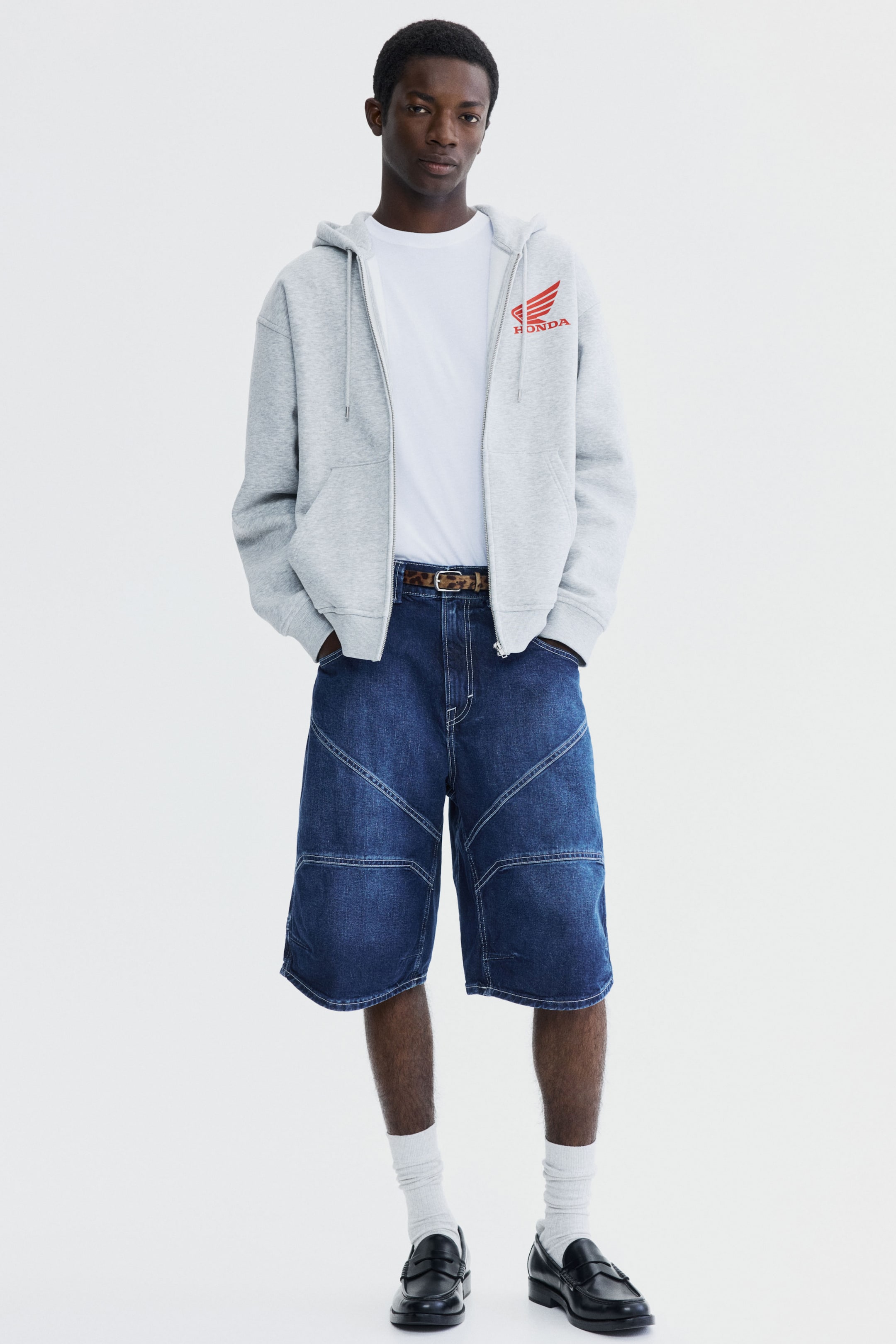 View larger image: Dark blue denim shorts feature a wide-leg cut with distinctive white stitching forming geometric panels on the front, extending to just above the knees. A leopard print belt threads through the belt loops.