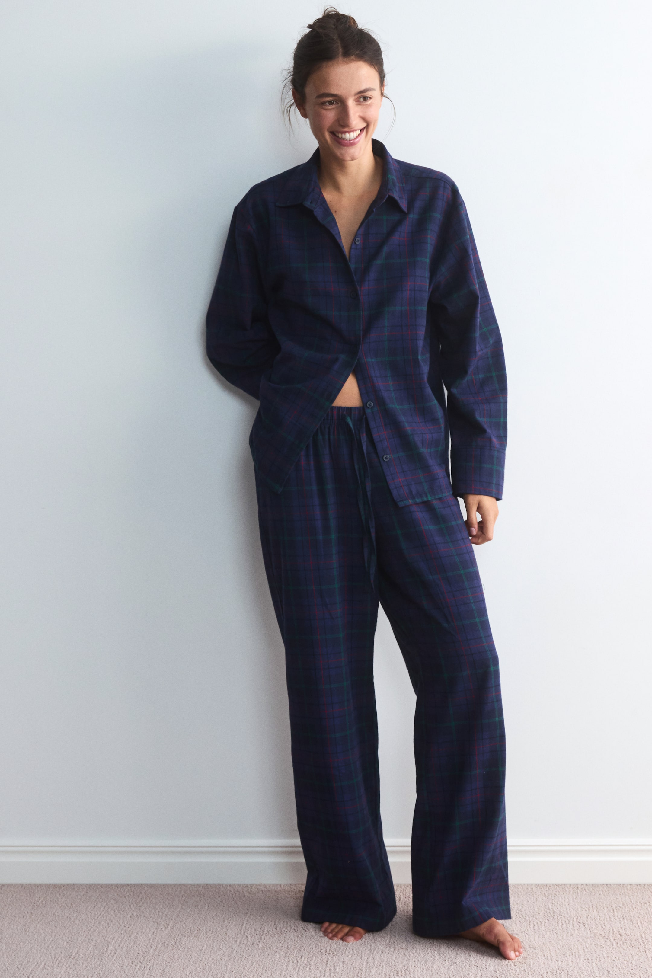 Größeres Bild anzeigen: Das Modell trägt ein dunkles, kariertes Flanell-Pyjama-Set, bestehend aus einem langärmeligen Hemd mit Kragen und Knopfleiste sowie einer dazu passenden Hose mit weitem Bein. Das Hemd ist unten teilweise aufgeknöpft und gibt den Blick auf den elastischen Bund mit Kordelzug der Hose frei.
