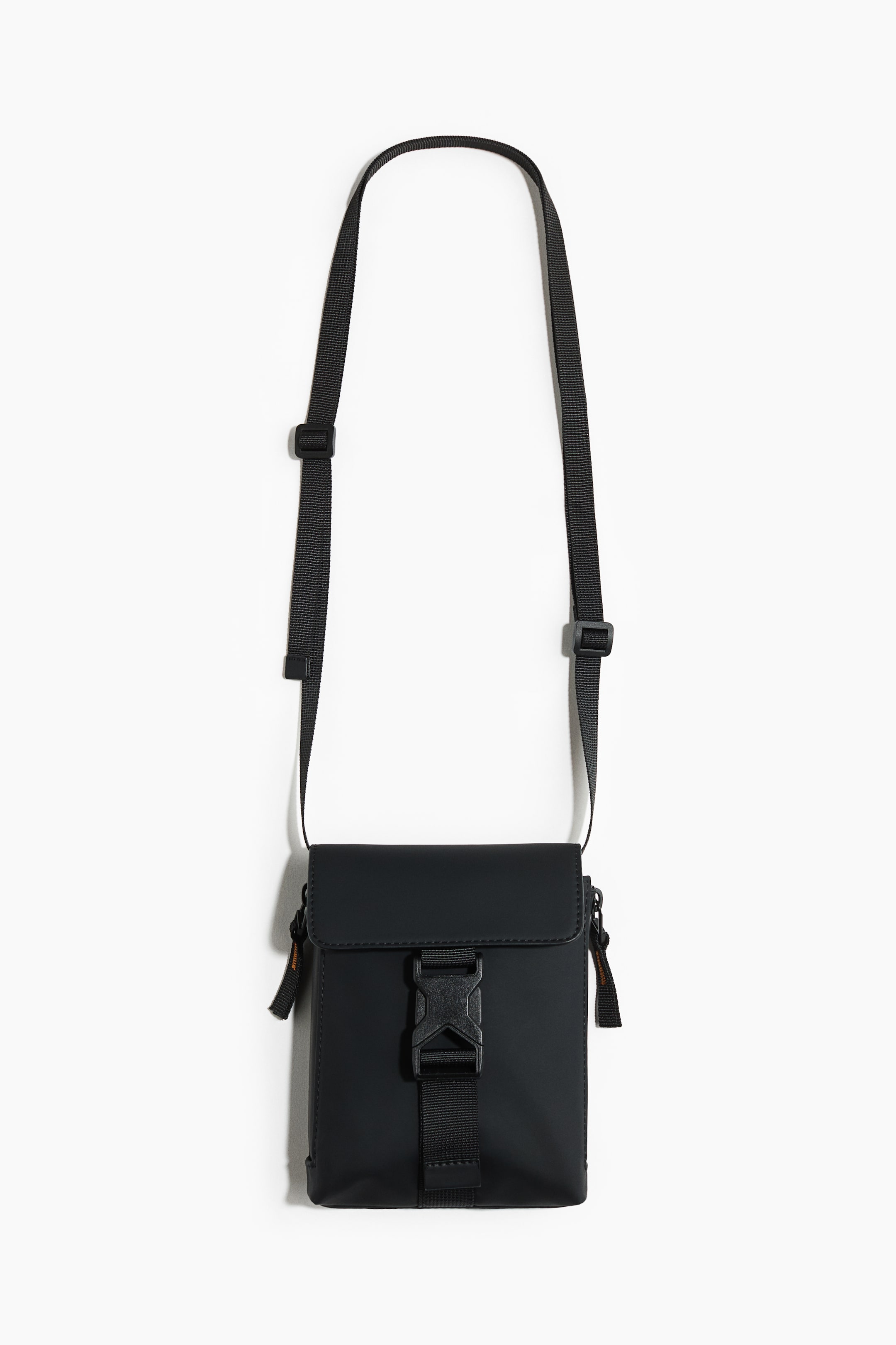 View larger image: A black cross-body bag is shown facing front, with a matte finish and a flap closure secured by a black plastic buckle on a vertical webbing strap. It features an adjustable black shoulder strap and small zipper pulls with orange accents on its sides.