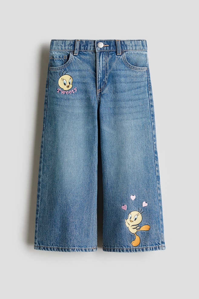 FRONT-Ansicht von blauen Baumwoll-Denim-5-Pocket-Jeans. Die Jeans zeichnen sich durch eine lockere Passform an Hüfte und Oberschenkel mit weiten Beinen aus und werden mit einem Reißverschluss und einem Druckknopf geschlossen. Ein Tweety Bird-Grafik mit dem Text 'Tweety' erscheint auf dem oberen linken Bein, während eine andere Tweety Bird-Grafik mit rosa Herzen auf dem unteren rechten Bein sichtbar ist. - 1