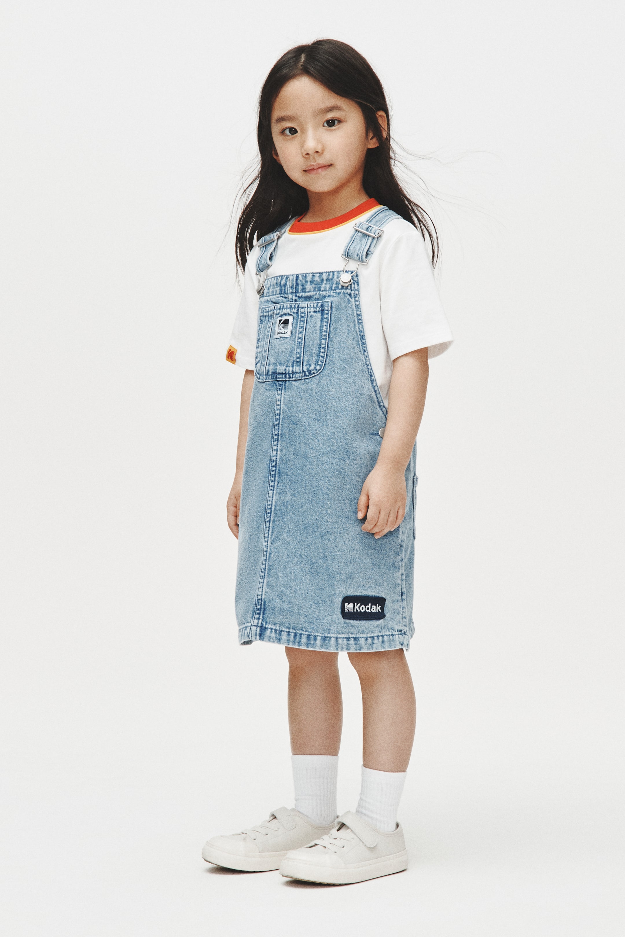 View larger image: A light blue denim overall dress is showcased from the front, featuring adjustable shoulder straps with metal hardware and a square bib detailed with a "Kodak" label on its patch pocket. An additional "Kodak" patch is visible on the lower front of the A-line skirt, and the dress is layered over a white t-shirt with an orange and yellow ribbed collar.