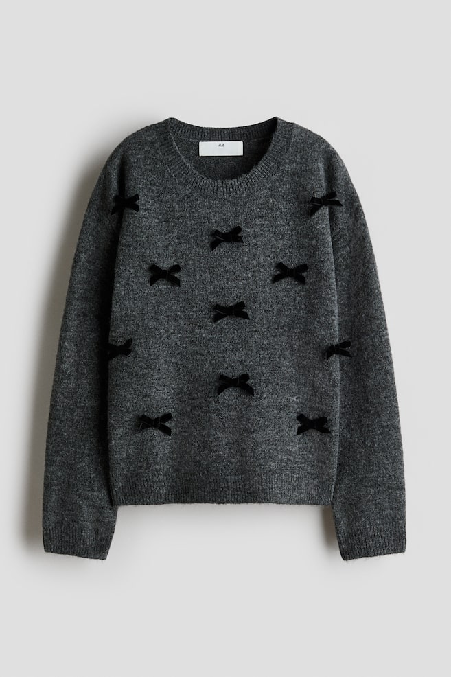 Ein dunkelgrauer Strickpullover ist von vorn zu sehen und verfÃ¼gt Ã¼ber einen gerippten Rundhalsausschnitt, Ã¼berschnittene Schultern sowie gerippte BÃ¼ndchen und einen gerippten Saum. Kleine schwarze Samtschleifen zieren die Vorderseite und die Ãrmel des Pullovers. - 1