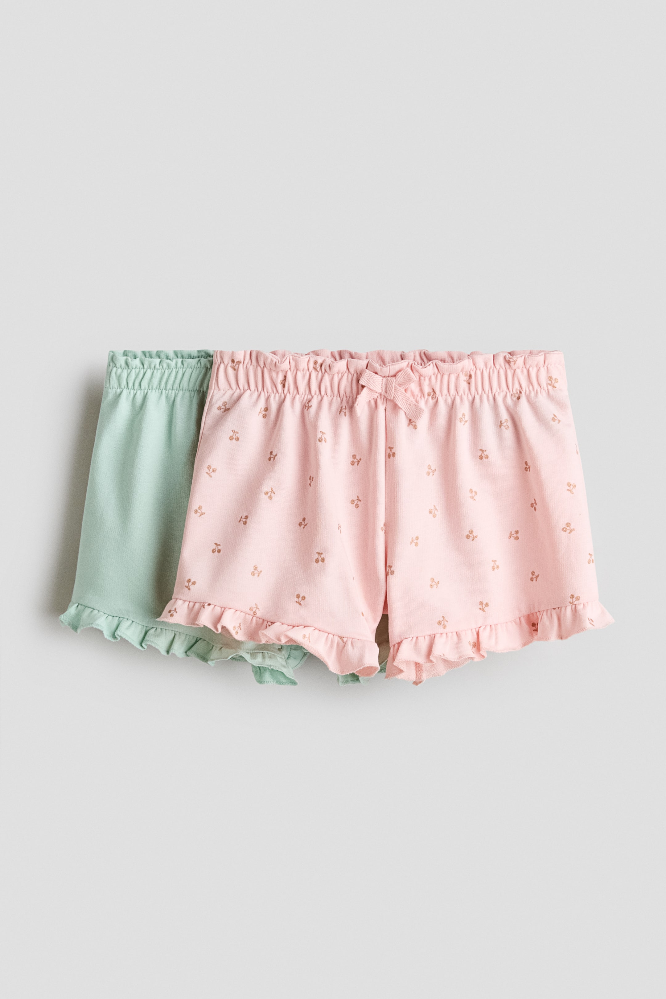 View larger image: "Makikita ang dalawang pares ng shorts mula sa harap na anggulo, kung saan ang isang pares na pink ay bahagyang nakapatong sa isang pares na mint green. Ang pink na shorts ay may maliit na pattern ng cherry at isang pandekorasyong laso sa ruched elastic waistband, habang parehong shorts ay may ruffled hems."