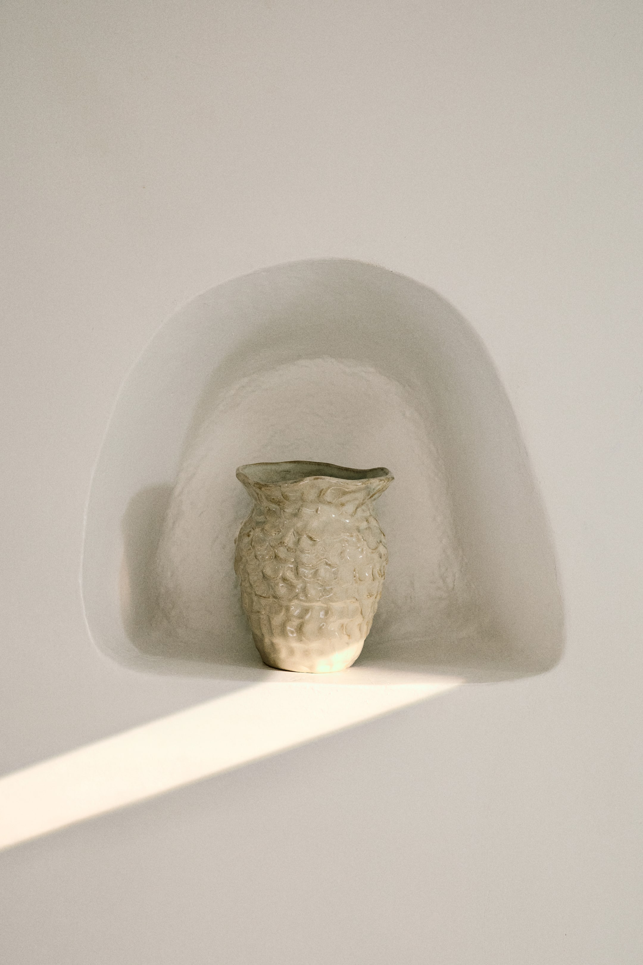 拡大画像を表示: 淡い色合いの花瓶が前面を向け、表面には不規則な隆起模様が施されています。胴体は下部に向かって細くなり、開口部は波状で有機的な縁を形成しています。