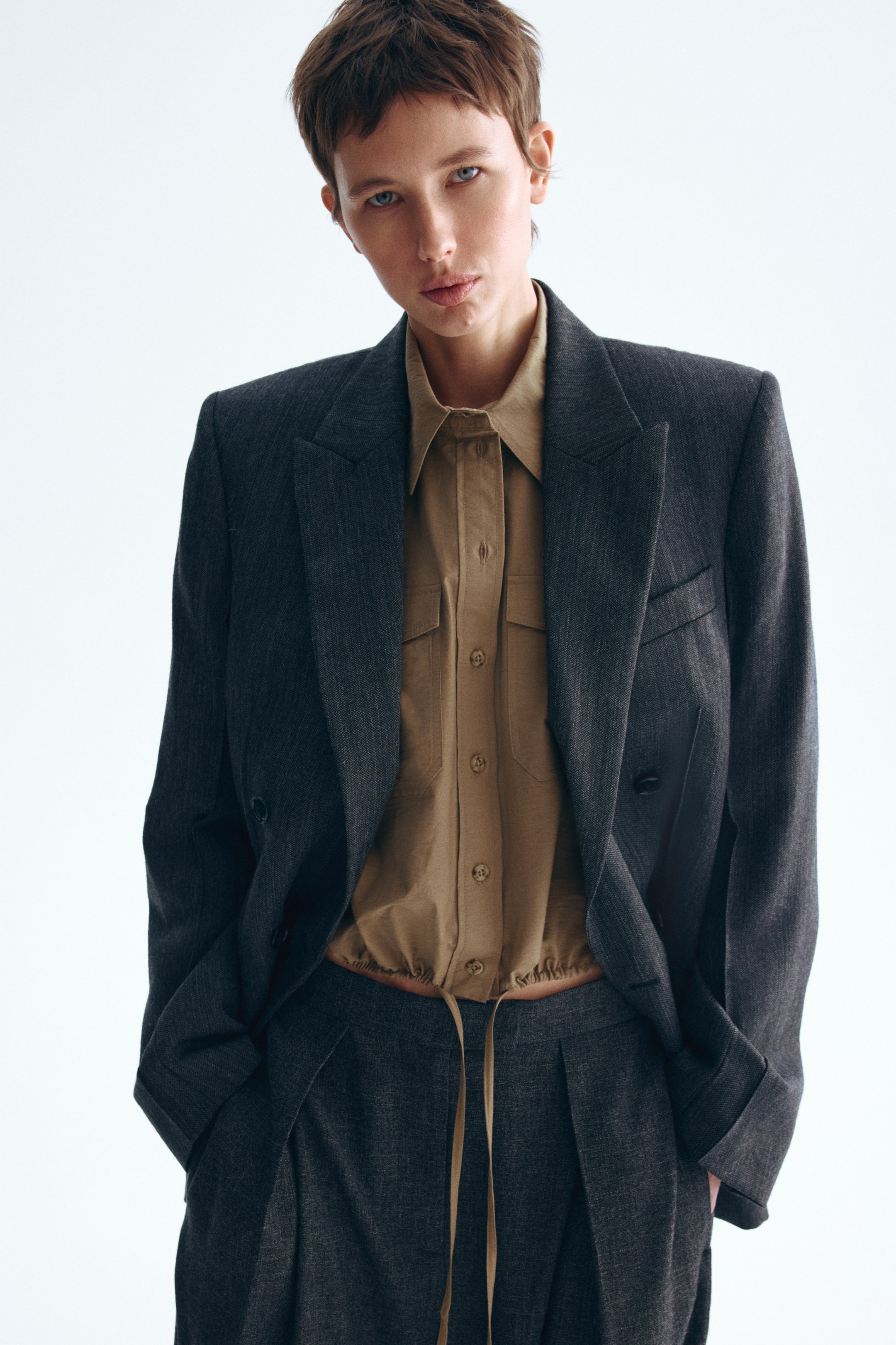 Visa större bild: En mörkgrå dubbelknäppt blazer med skårade slag och klaffickor, bärs öppen över en beige skjorta med krage. Skjortan har två bröstfickor och ett samlat dragsnöre i midjan, och den syns ovanför matchande mörkgrå plisserade byxor som också visar ett synligt dragsnöre.