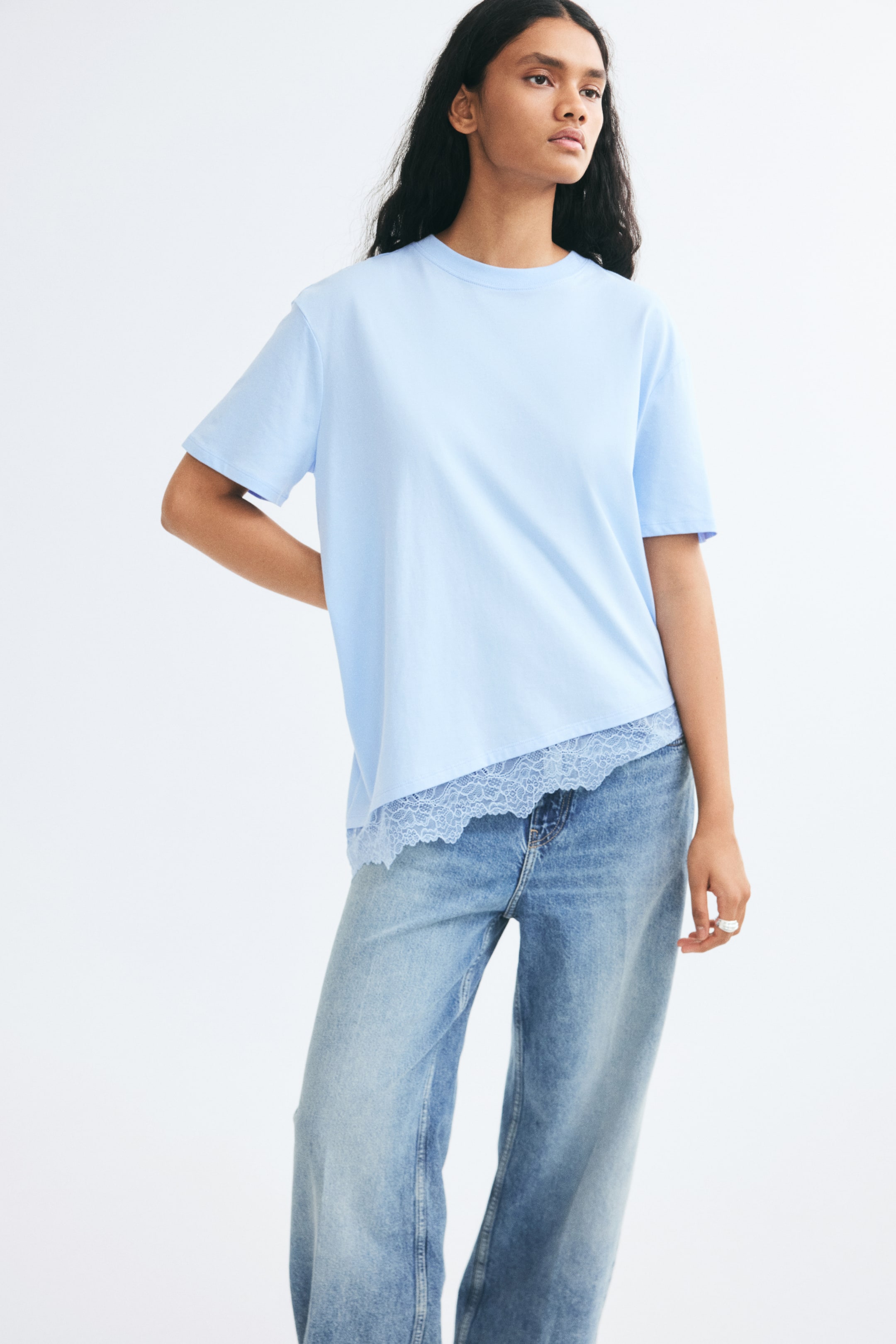 View larger image: A light blue, relaxed-fit T-shirt is worn by the model, showcasing a ribbed crew neck, dropped shoulders, and an asymmetrical hem with scalloped lace trim. The T-shirt is styled over light-wash, wide-leg denim jeans.