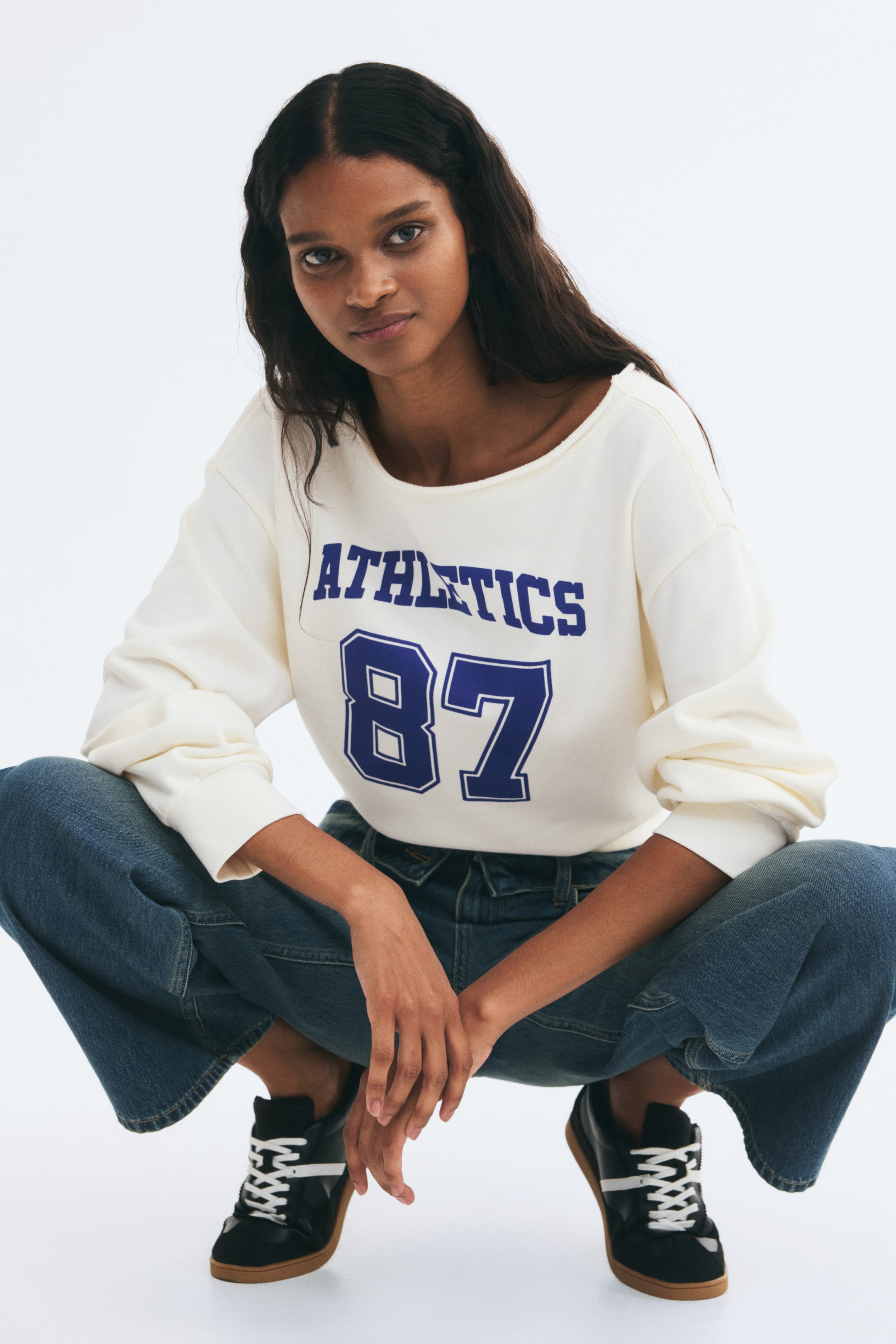 View larger image: A loose-fit, off-the-shoulder cream sweater featuring a blue "ATHLETICS 87" print on the chest, dropped shoulders, and ribbed cuffs is worn by the model. The sweater is styled with blue jeans and black sneakers.