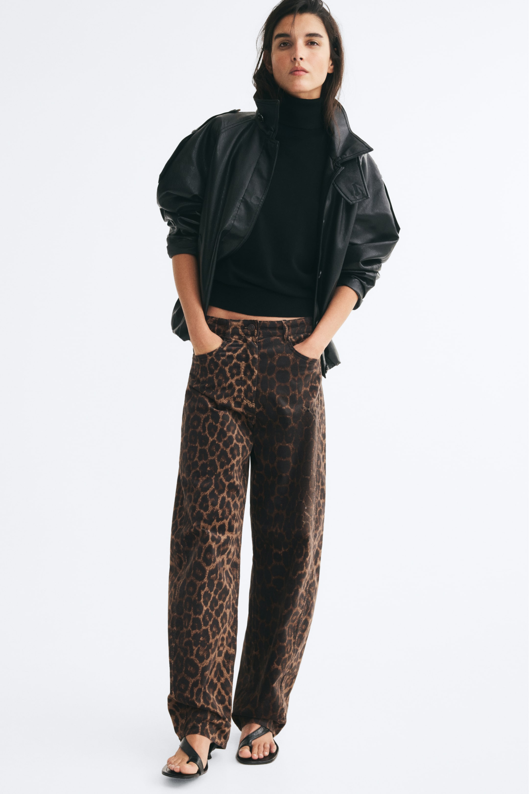 View larger image: High-waisted, five-pocket jeans featuring a brown and black leopard-print pattern and curved, voluminous legs are worn by the model, who is standing with her hands in the front pockets. The jeans are paired with a black roll-neck top, an open black leather jacket, and black flat sandals.