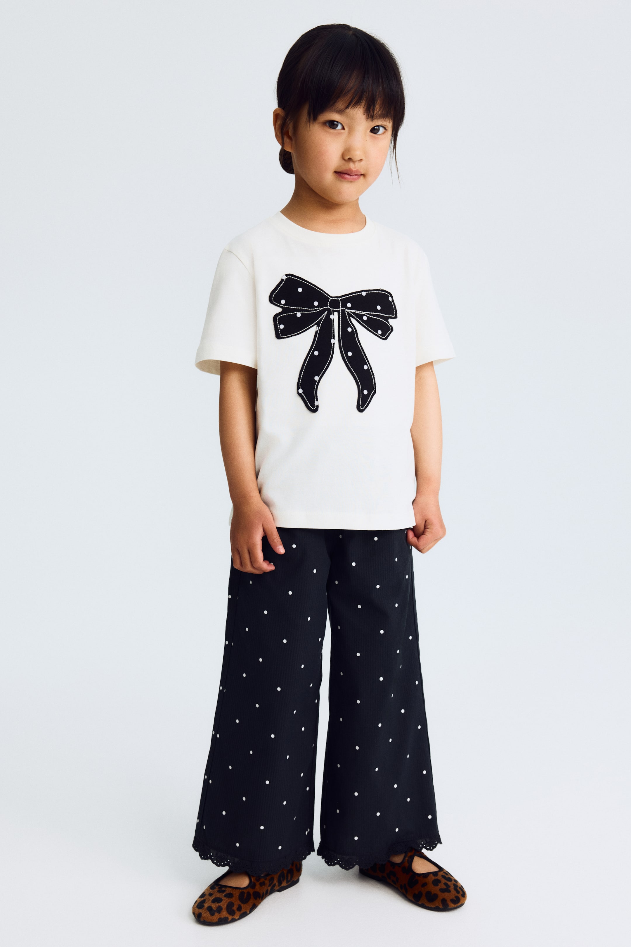 View larger image: An off-white short-sleeved t-shirt showcases a prominent black bow appliqué adorned with white polka dots and white stitching on the front. It's paired with dark, wide-leg ribbed pants featuring white polka dots and a black scalloped lace trim at the hem, complemented by leopard print ballet flats.